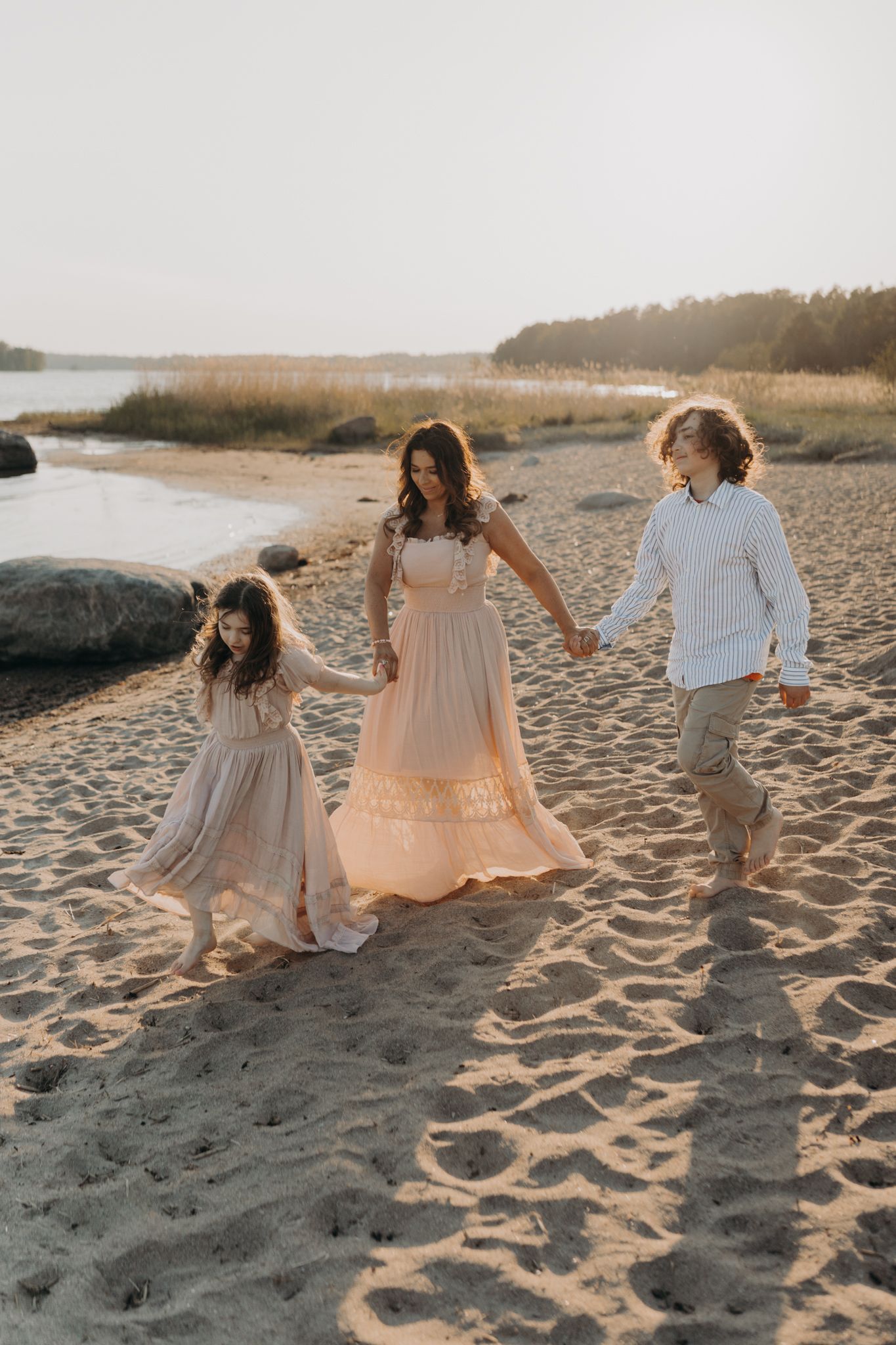 A mother and two children hold hands walking along a sandy beach at golden hour.