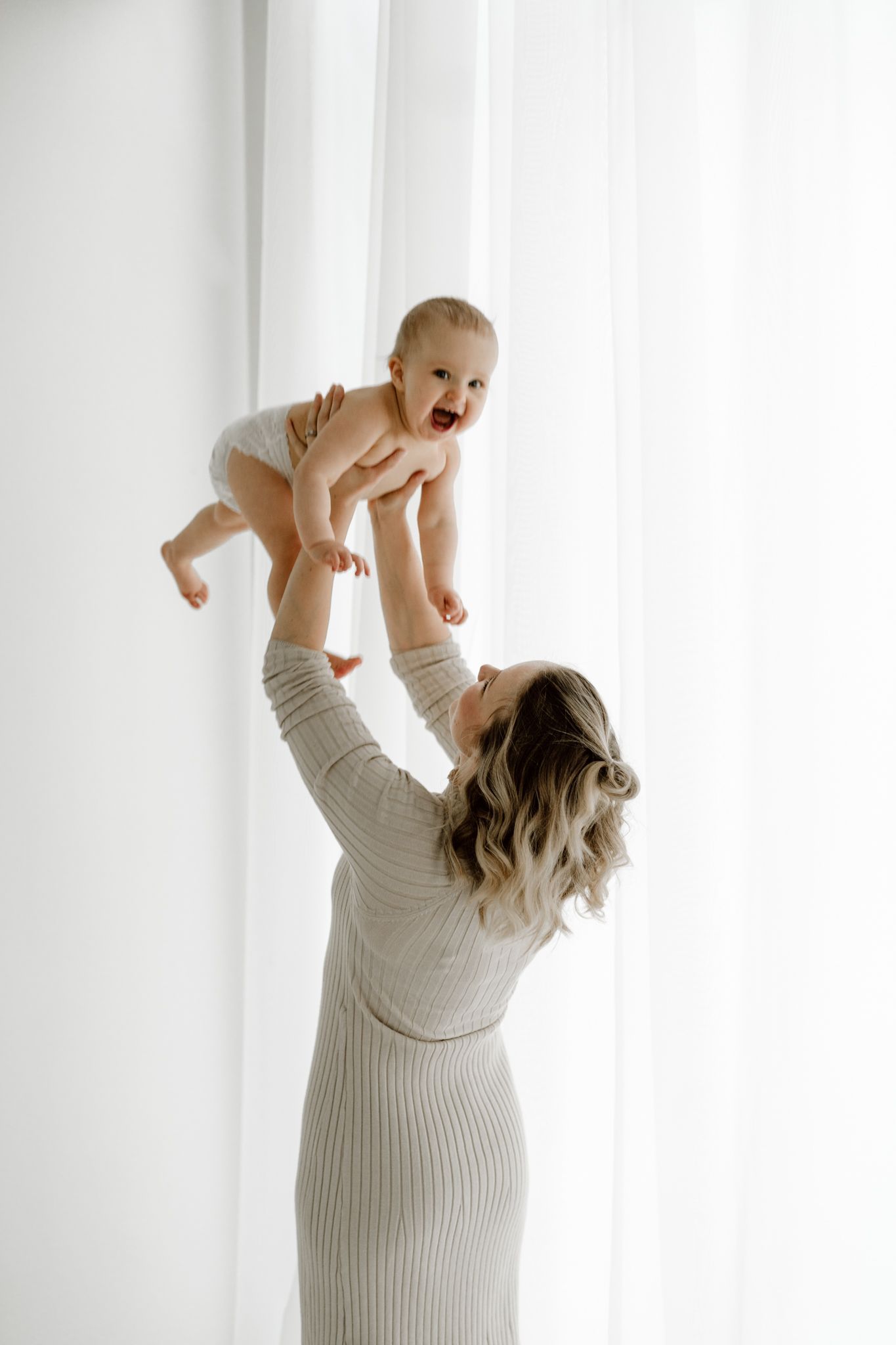 Mother in ribbed dress lifting giggling baby in diaper toward white curtained window.