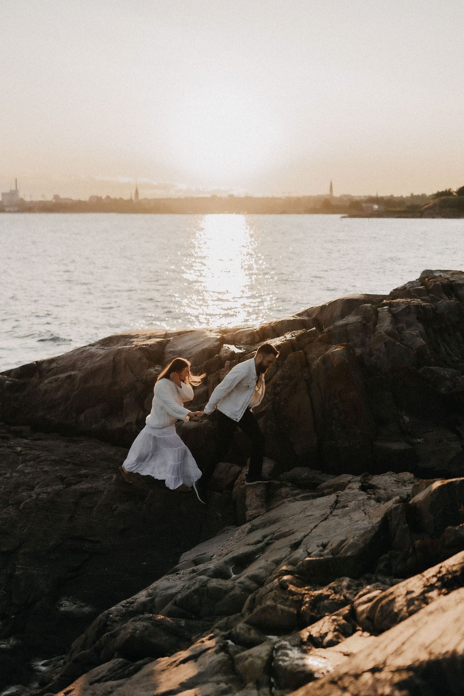Couple climbing sun-drenched coastal rocks with a glowing sunset reflecting over calm water.
