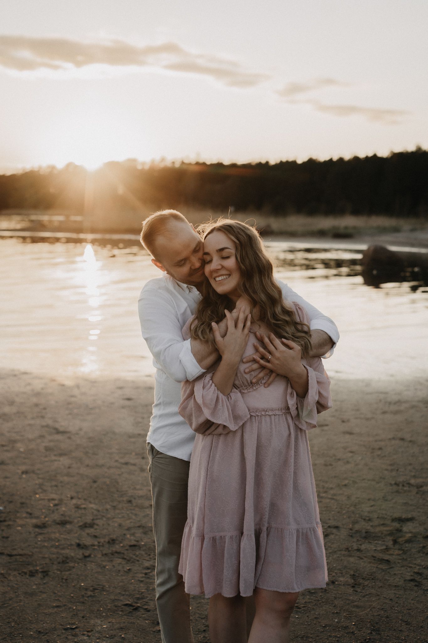 Man tenderly embraces woman in pink dress from behind on a sandy beach during a glowing sunset.