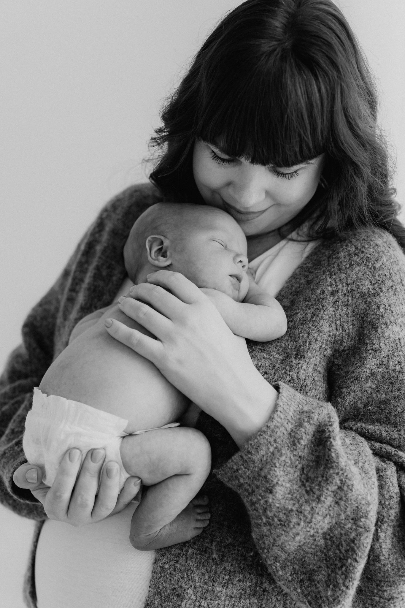 Black and white photo of a mother in a gray sweater tenderly holding her sleeping newborn baby.