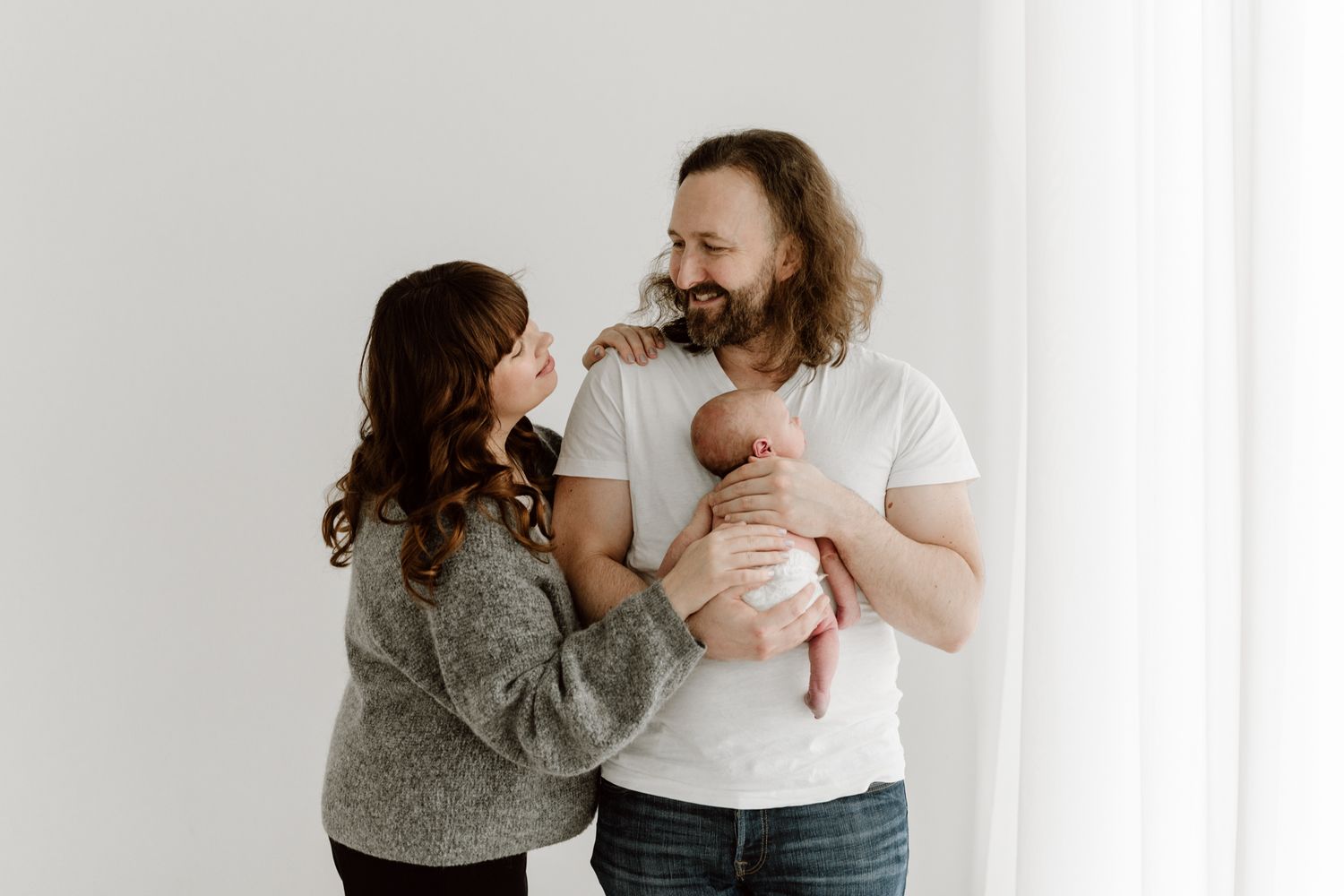 Happy parents smiling at each other while holding their newborn baby in a bright studio.