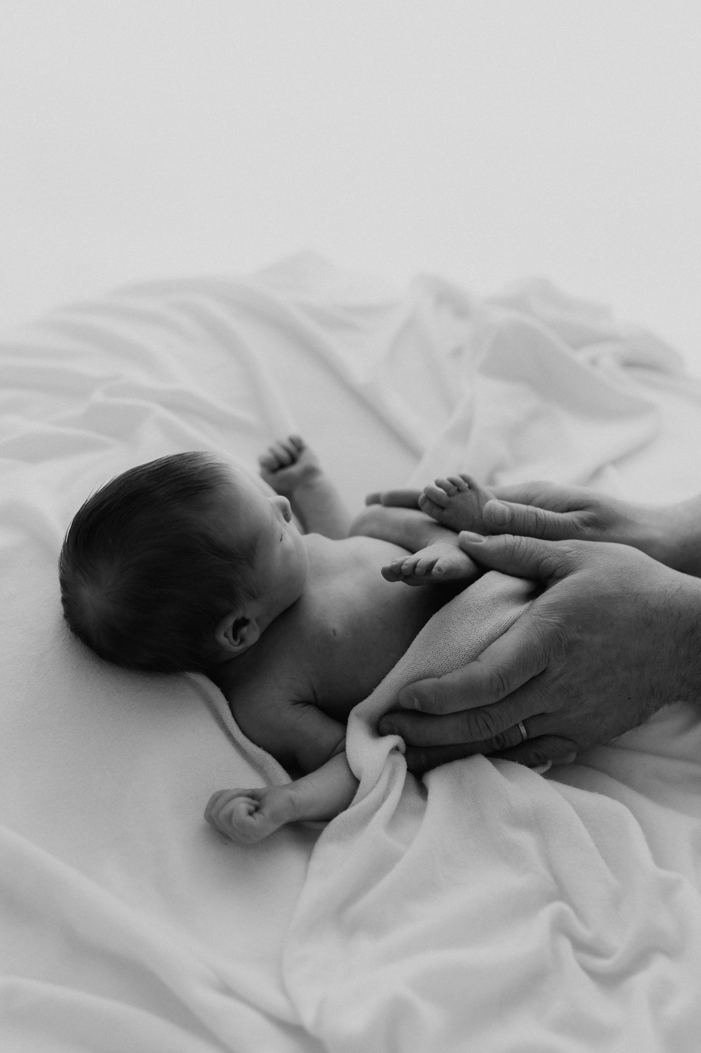 Black and white photo of newborn baby lying on white blanket with parent's hands tenderly cradling the infant.