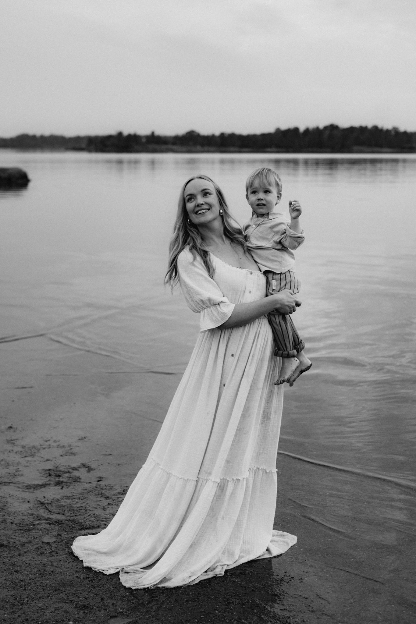 Black and white photo of a smiling mother in a white maxi dress holding her toddler by a lake.