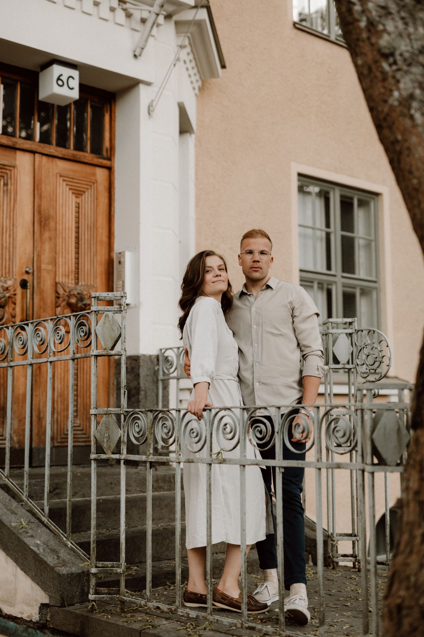 Elegant couple posing by ornate iron gate in front of a pink building with wooden door 6C.