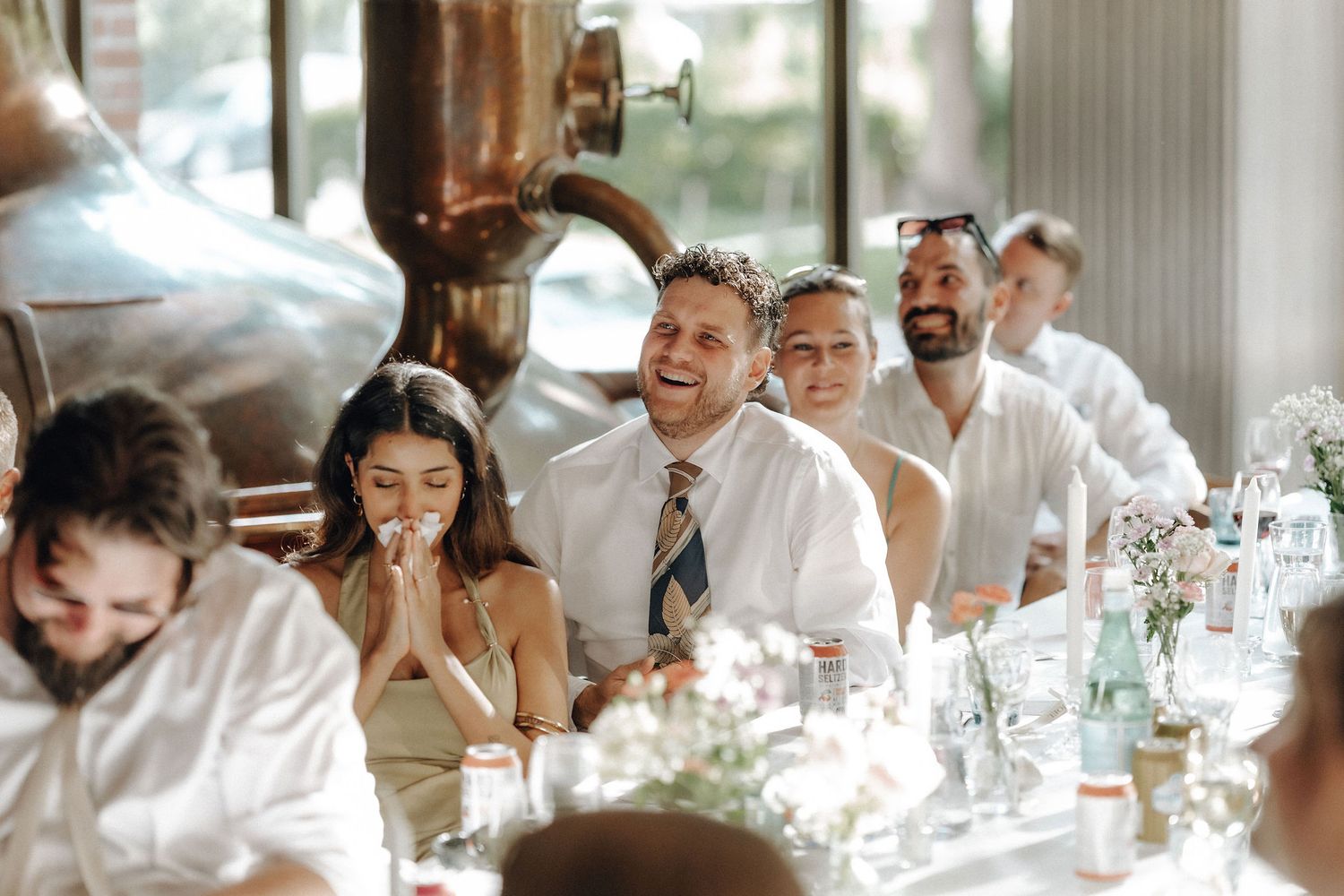 Guests laughing and enjoying wedding reception dinner at a brewery venue with copper tanks.