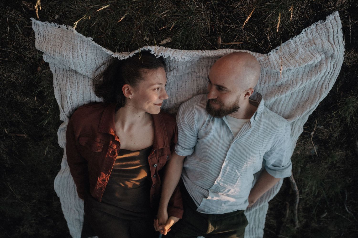 Couple lying on white blanket in grass, gazing at each other in an intimate moment.