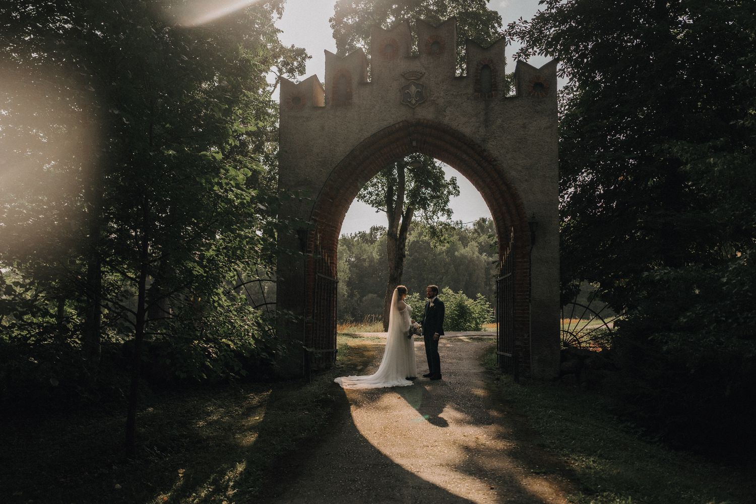 Bride and groom standing under stone arch gateway in sunlit forest wedding portrait.