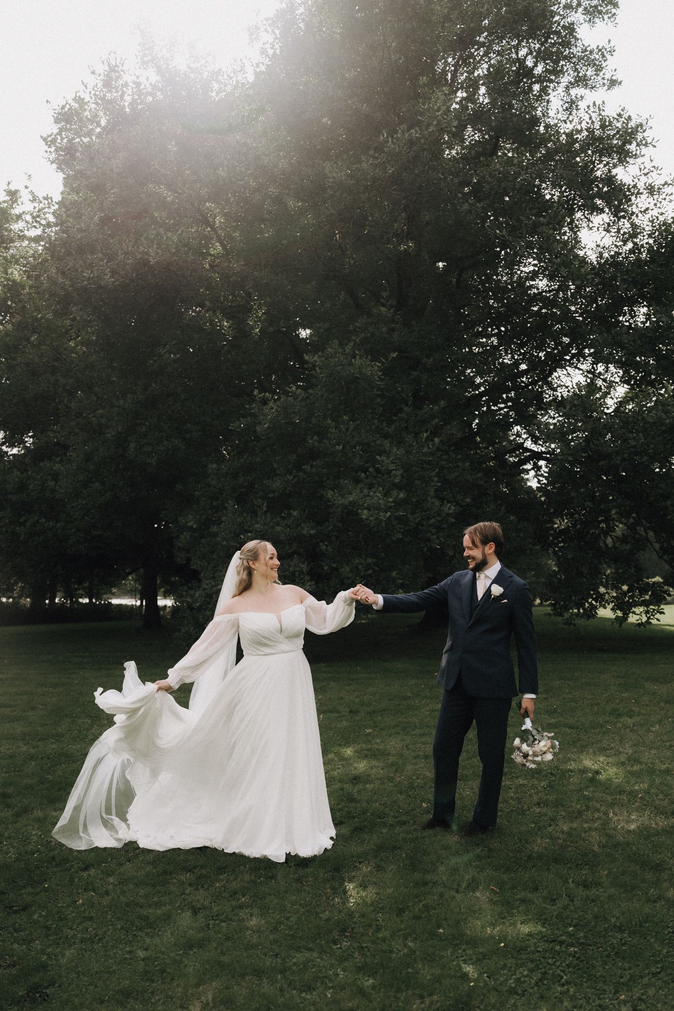 Bride and groom hold hands dancing in a sunlit green park, bride's white gown flowing in the breeze.