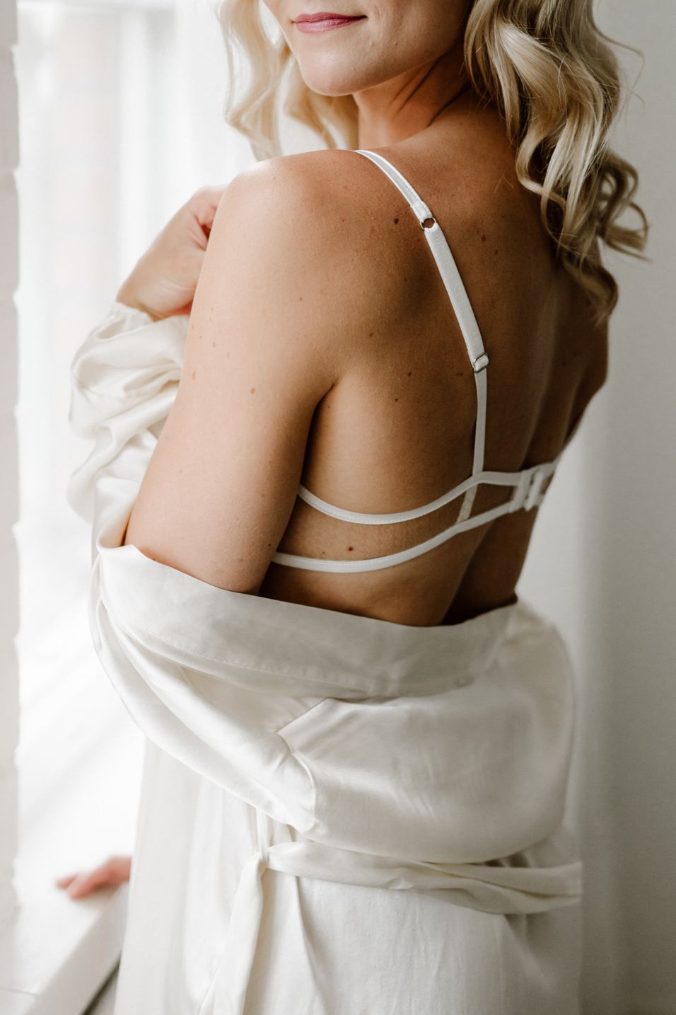 Woman in white robe revealing white bra straps on her back near a bright window.