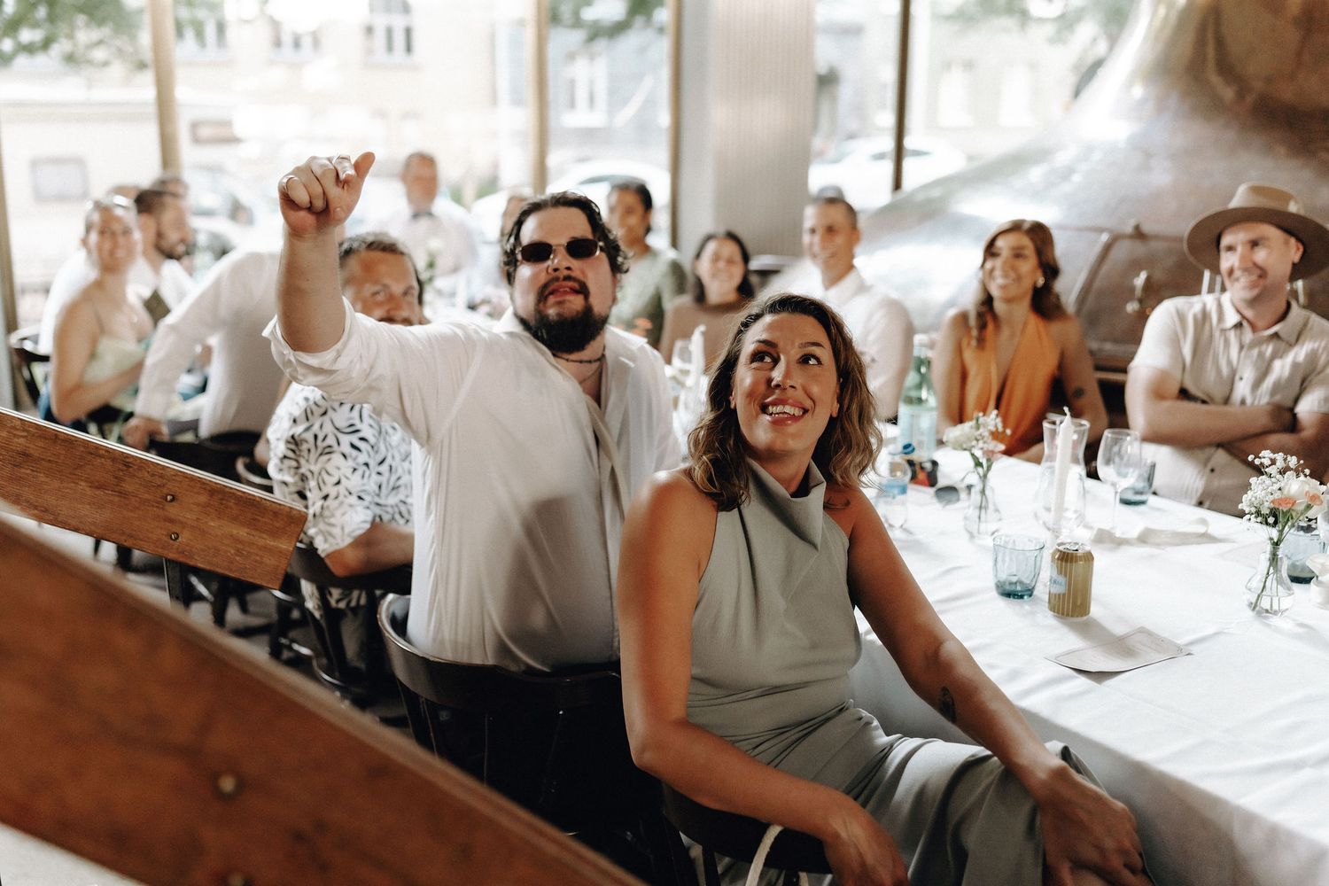 Wedding guests laughing and pointing during reception speeches at rustic brewery venue.