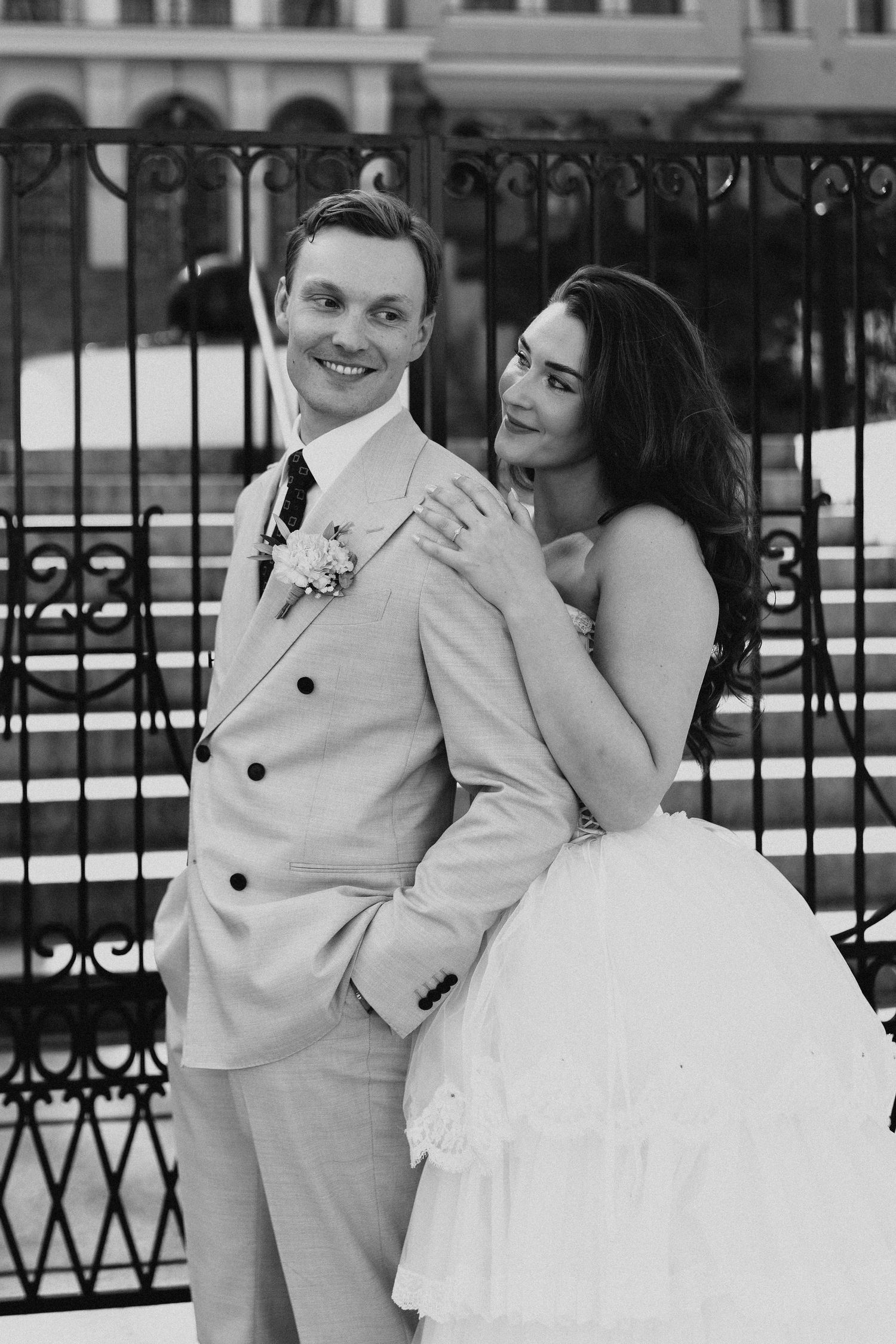 Black and white portrait of smiling groom in beige suit with bride embracing him from behind at iron fence.