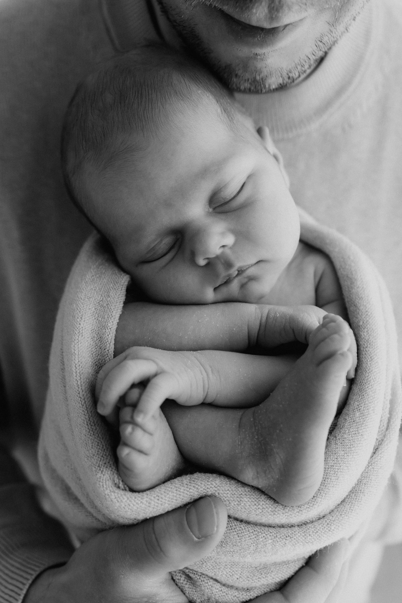Black and white photo of a sleeping newborn baby swaddled in a soft blanket, cradled in fathers hands.