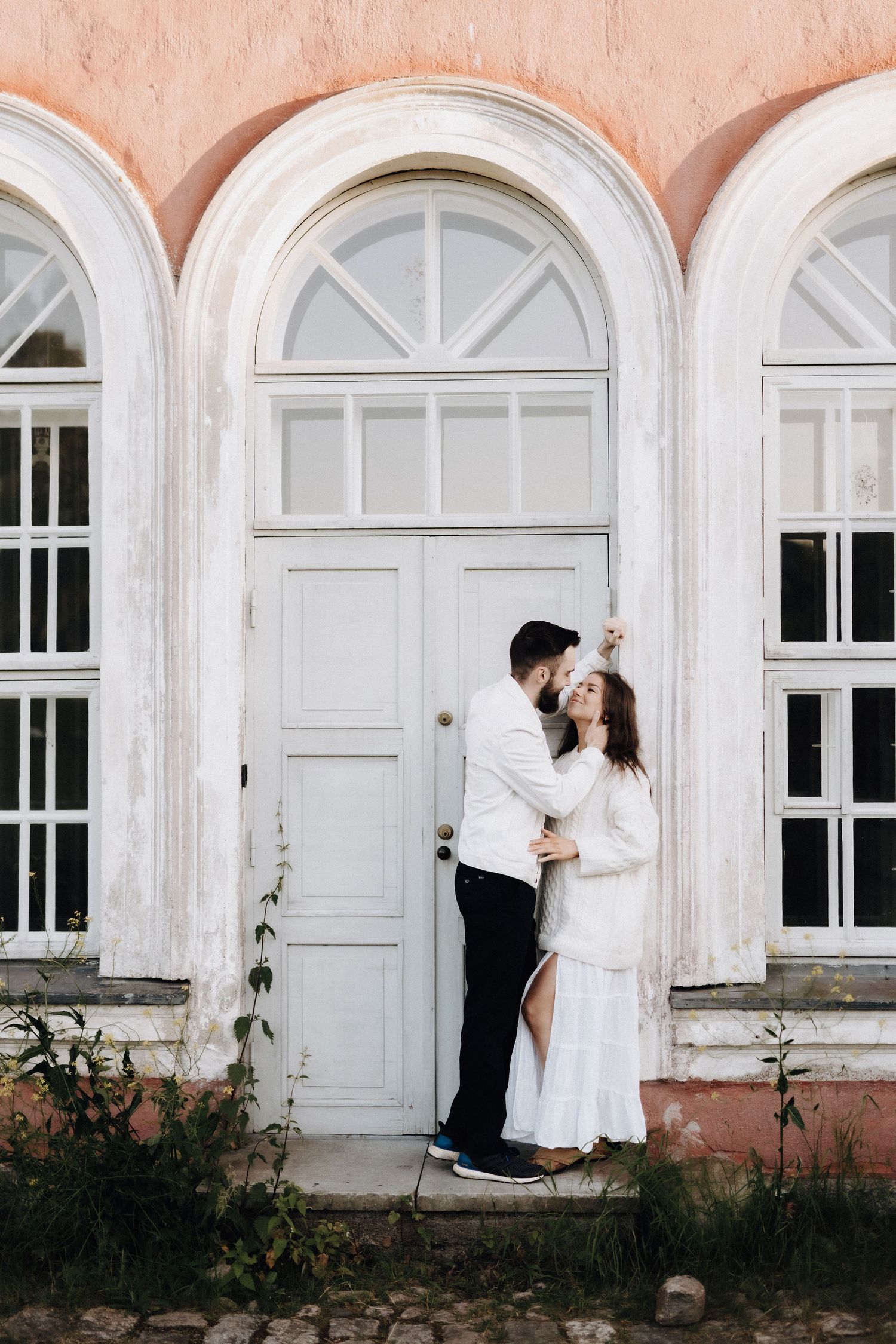 Romantic couple almost kissing in front of vintage arched white door on pink building.