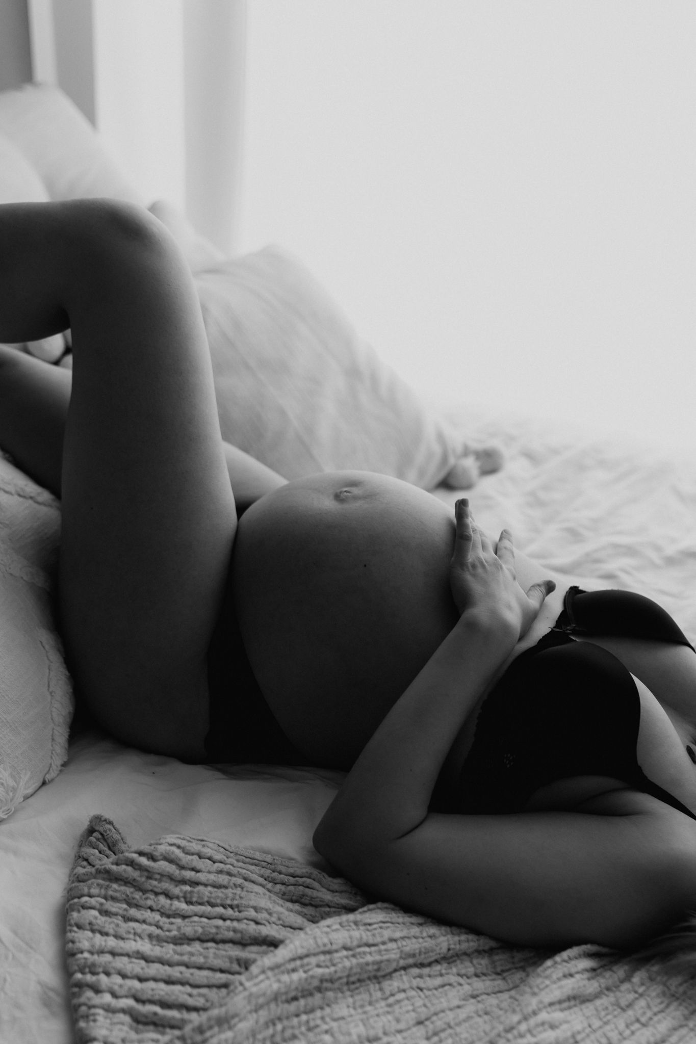 Pregnant woman in black lingerie lying on bed cradling her bare bump, intimate black and white maternity photo.