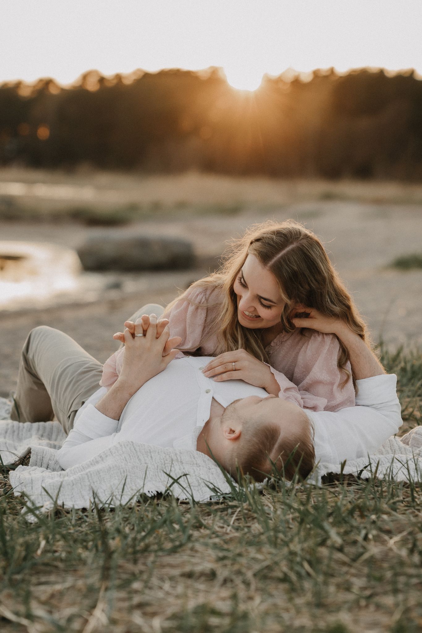 Romantic couple lying on blanket outdoors at golden sunset near waterfront, holding hands tenderly.