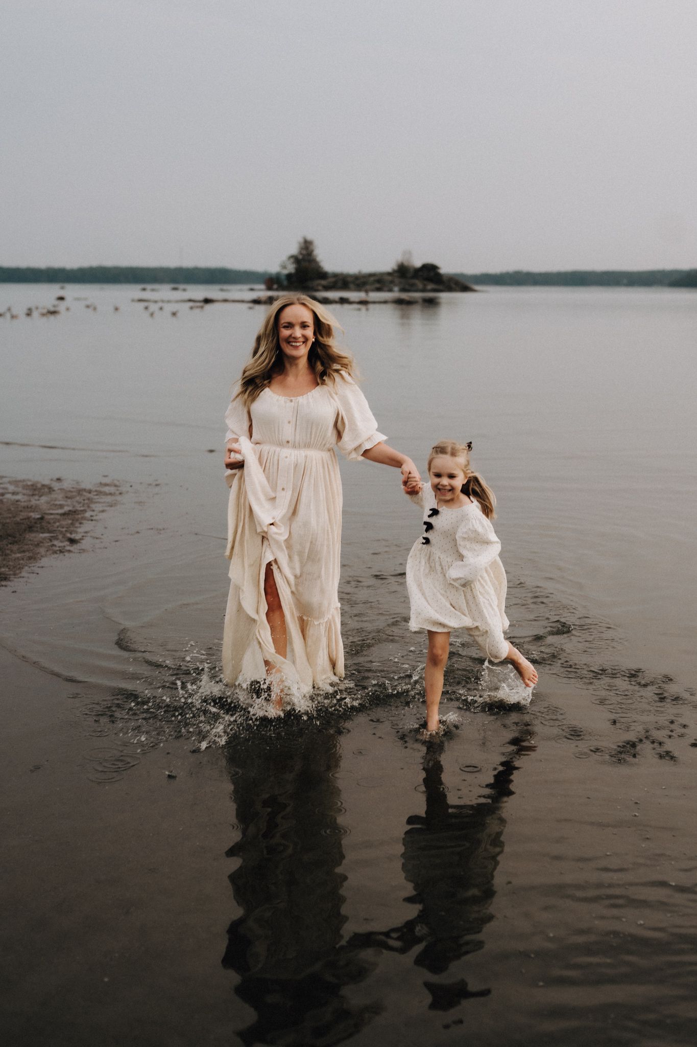 Mother and young daughter in white dresses joyfully splashing through shallow water on a lake beach.