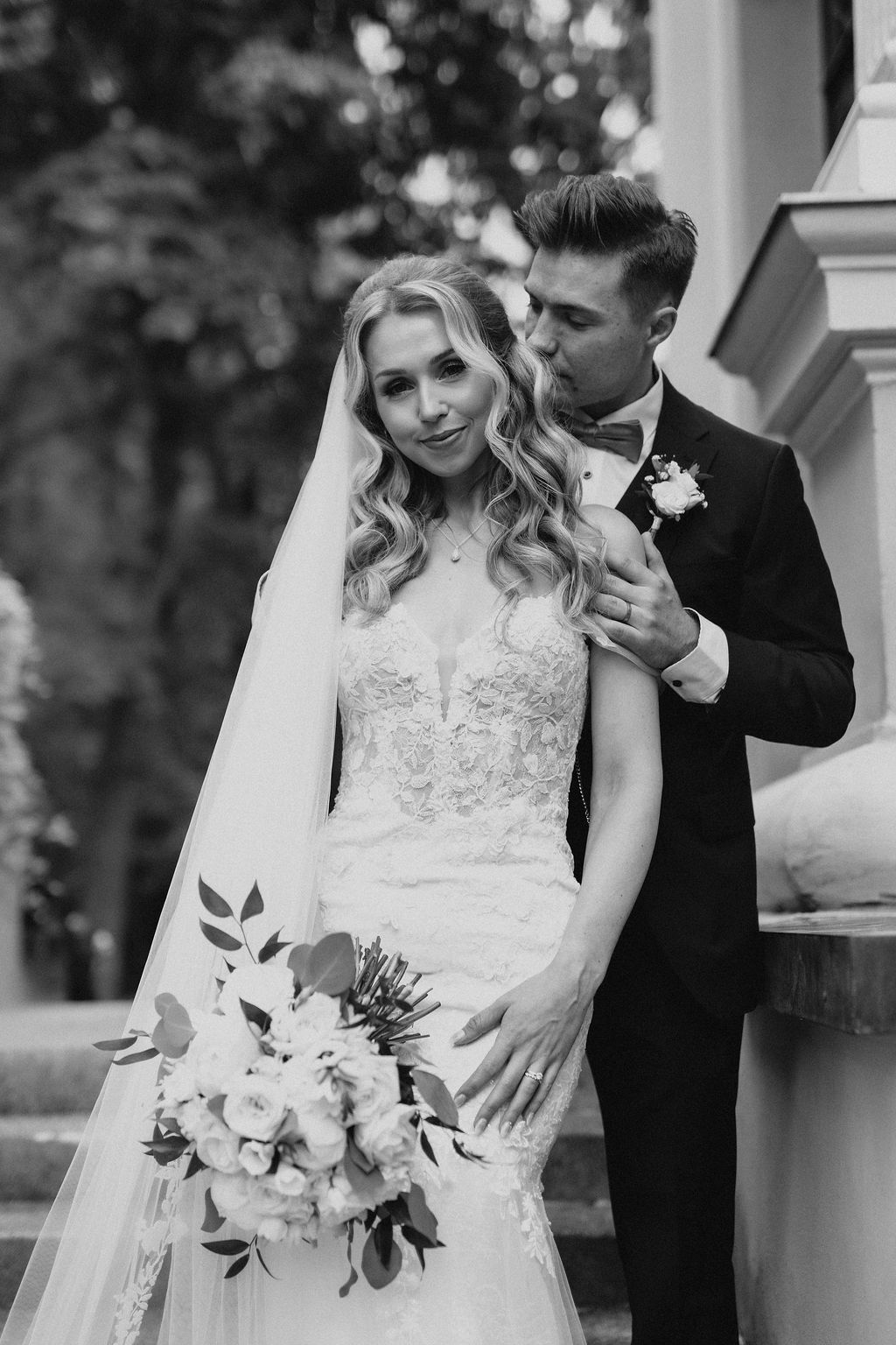 Black and white photo of groom kissing brides cheek as she smiles holding white floral bouquet.