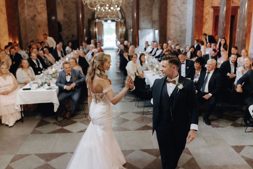 Bride and groom first dance in elegant chandelier-lit ballroom with applauding wedding guests.