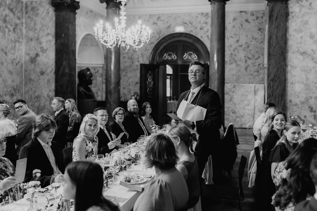 Wedding guest delivers heartfelt speech at formal reception dinner in grand ballroom with chandelier.