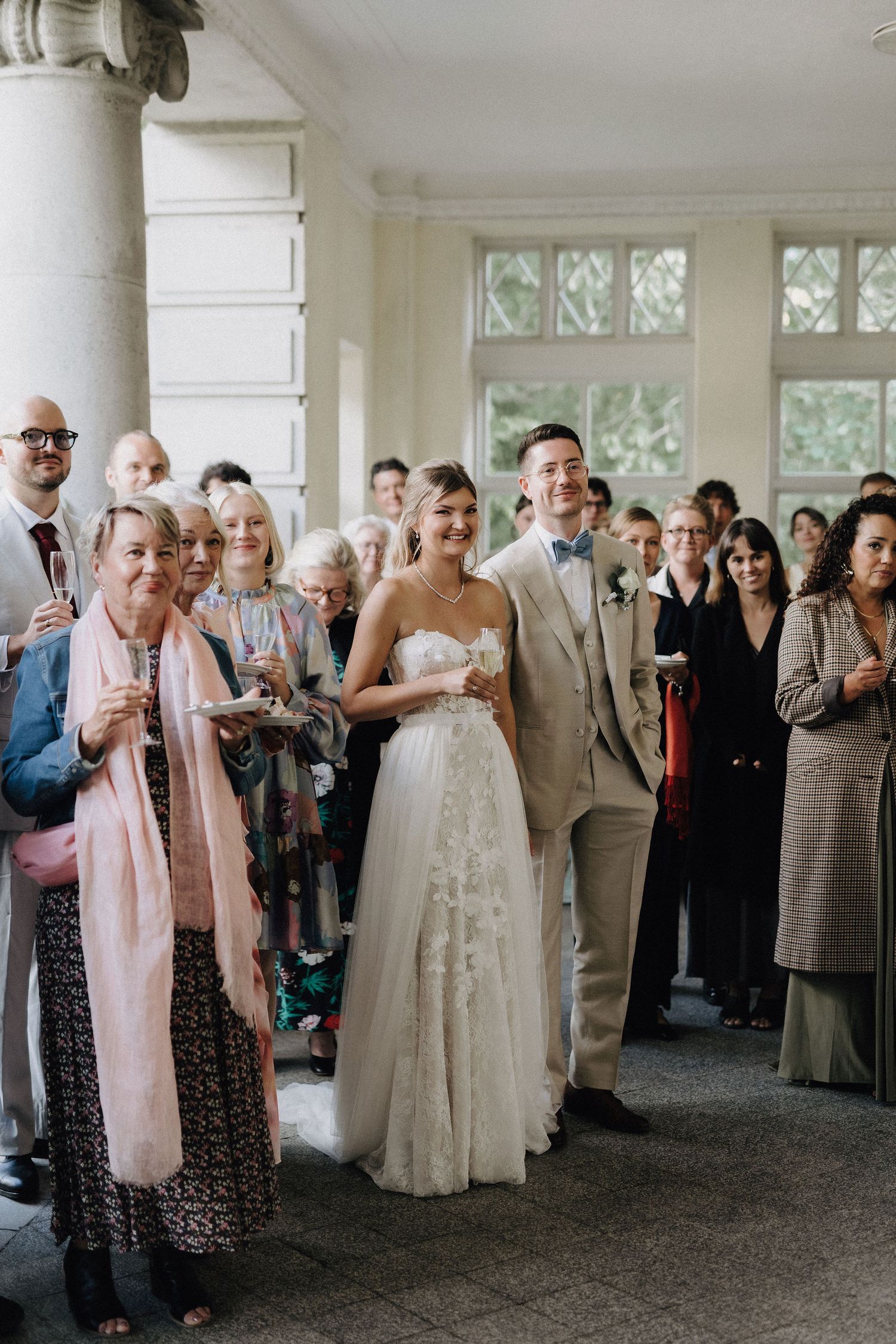 Bride in lace gown and groom in beige suit smile with wedding guests in elegant venue.