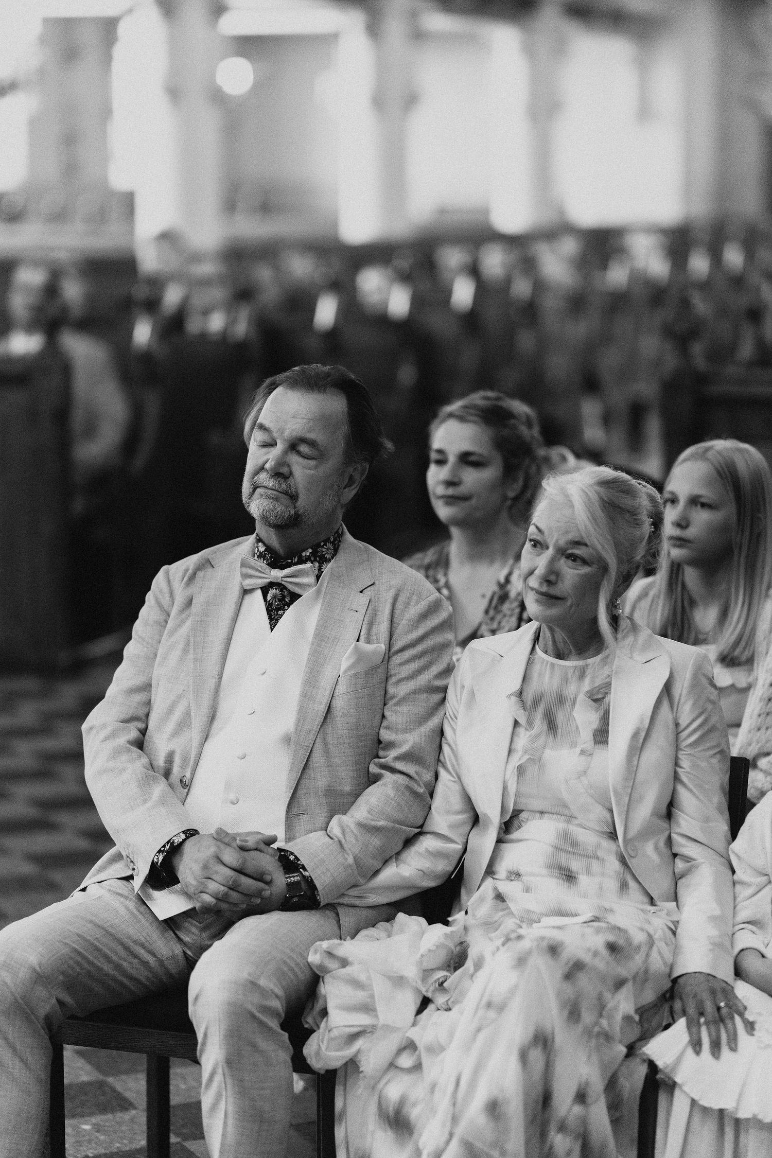 Black and white photo of wedding guests seated in church pews, including an older couple in formal attire.