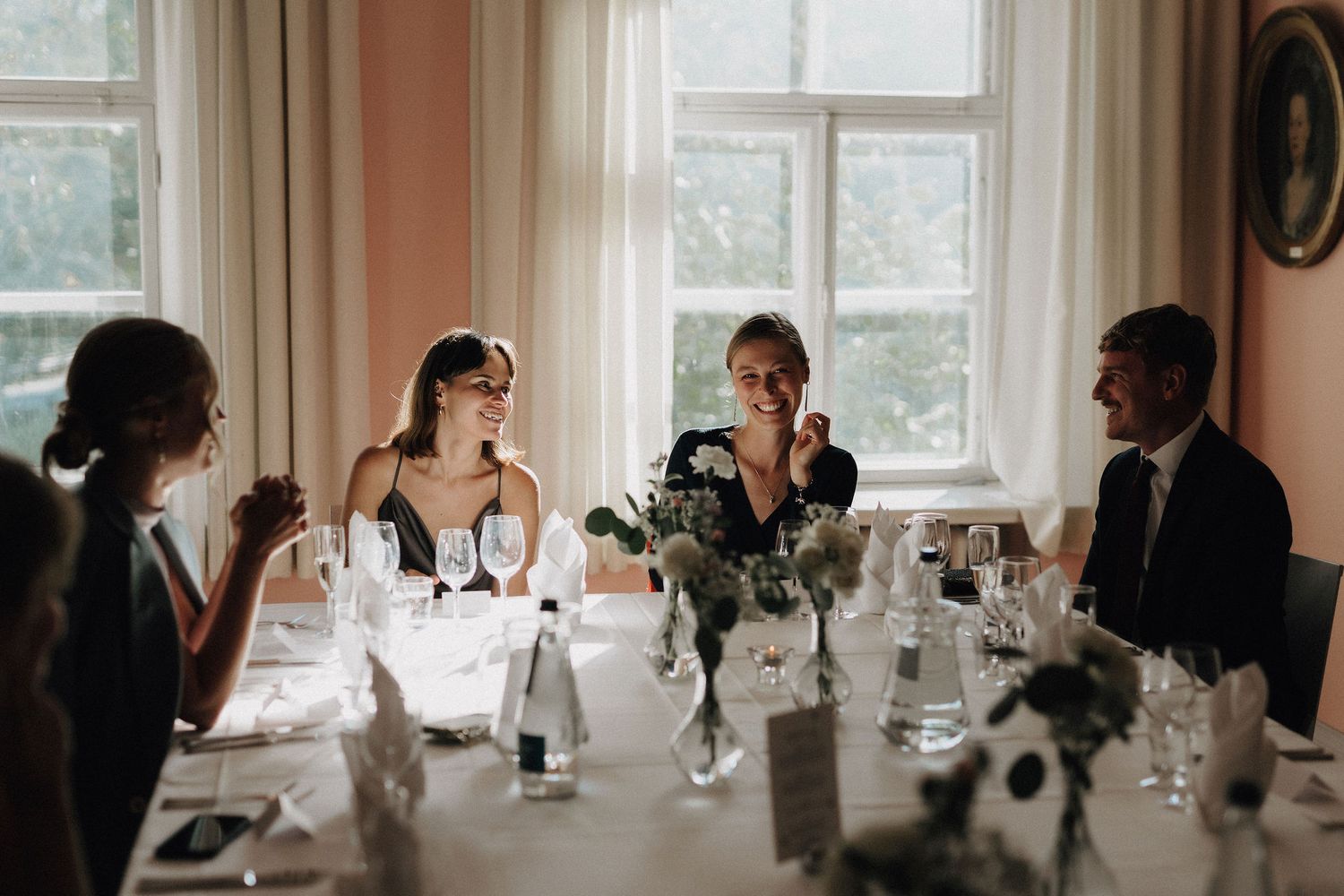 Wedding guests laughing and conversing at elegantly set dining table with floral centerpieces near bright windows.