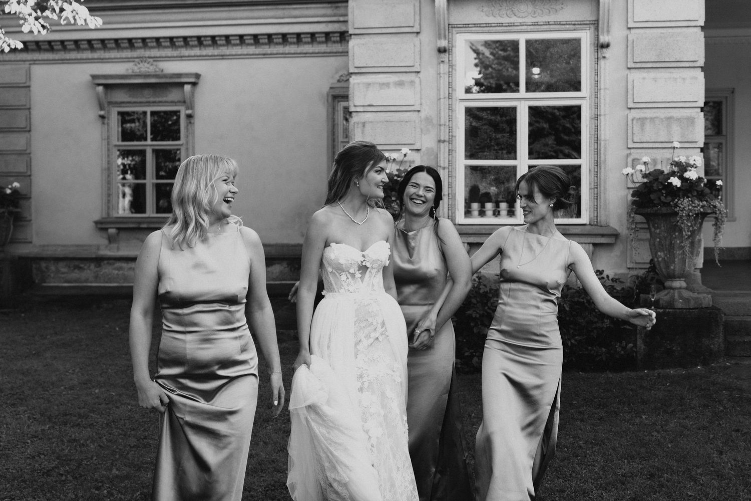 Bride and three bridesmaids in satin dresses walking and laughing together on lawn outside elegant venue.