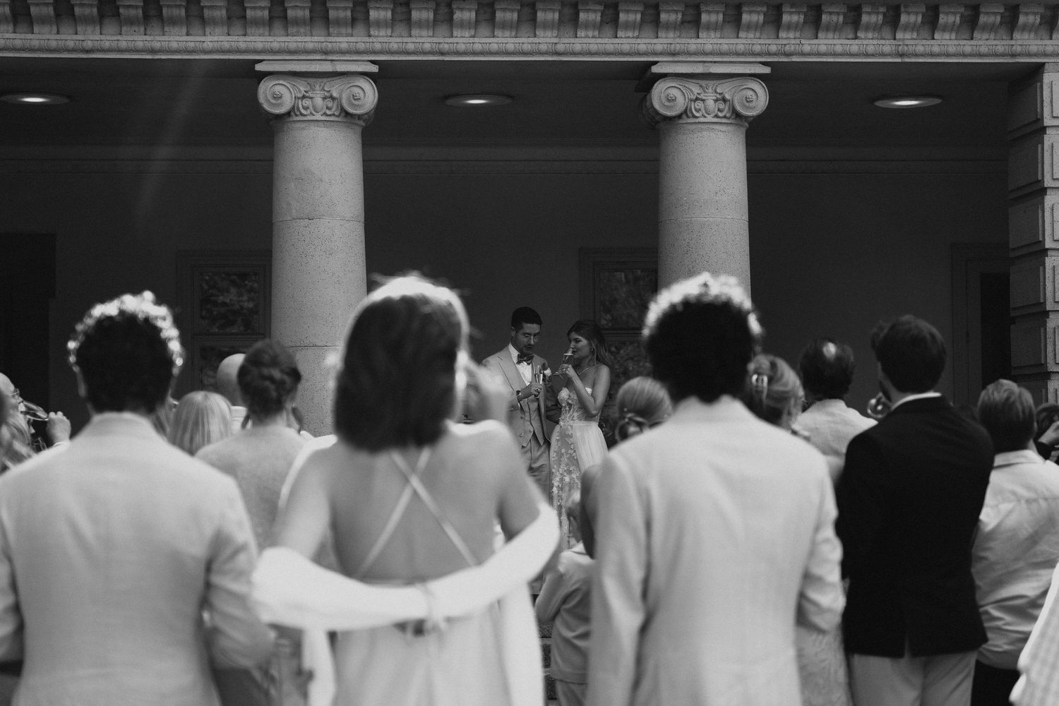 Wedding guests gathered before columned venue entrance watching outdoor ceremony, black and white.