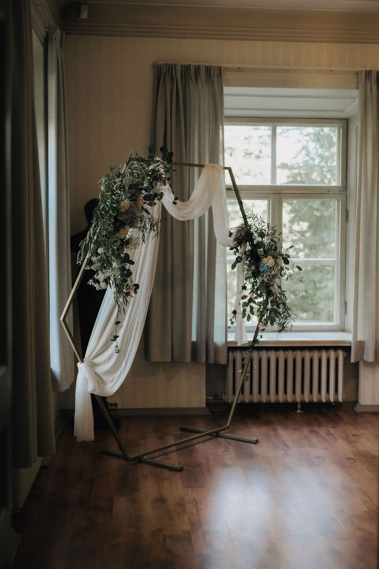 Elegant wedding ceremony arch decorated with white draping and greenery floral arrangements beside a window.