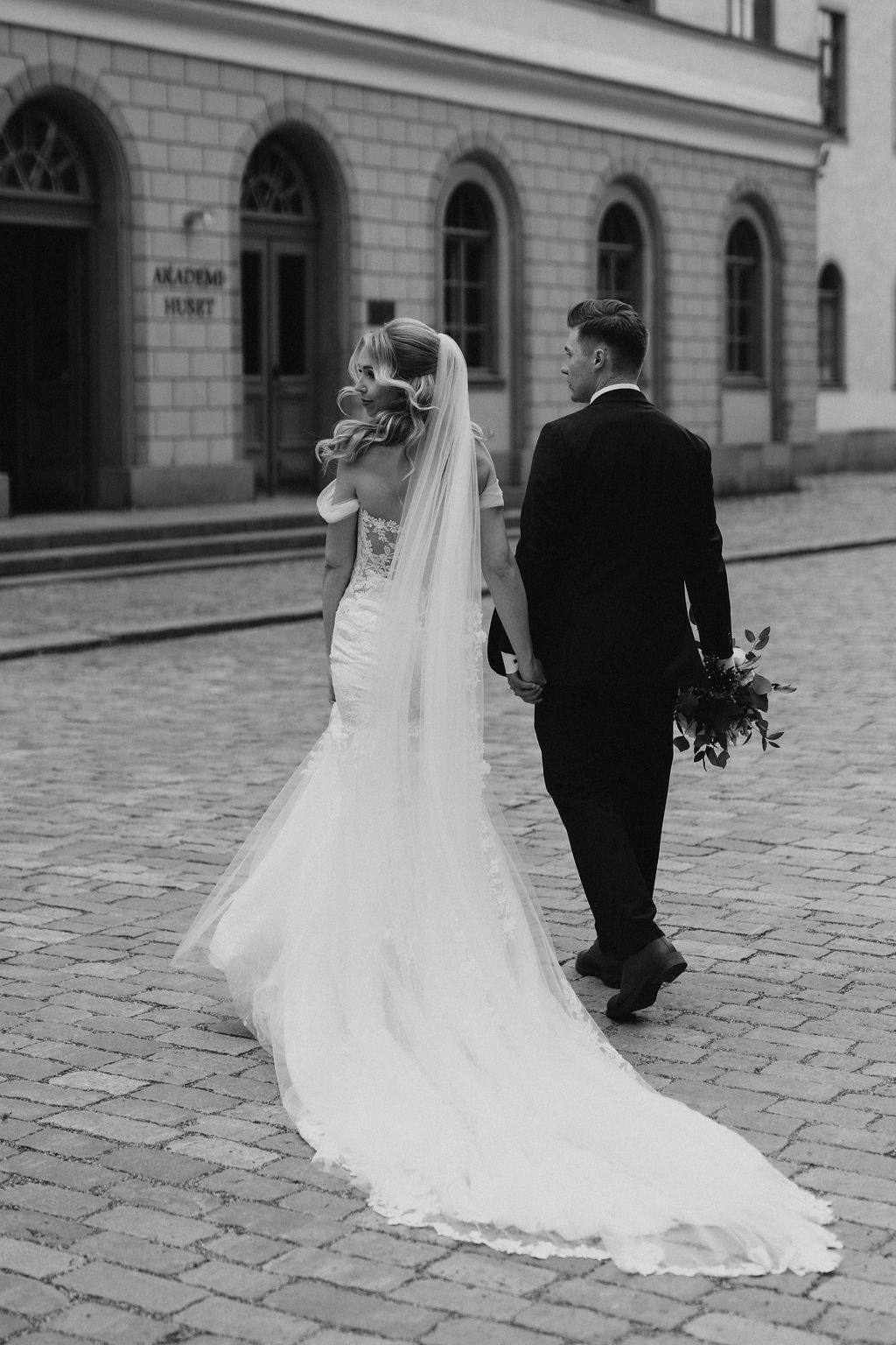 Black and white photo of bride and groom walking hand in hand on cobblestone street near Akademo Huset.