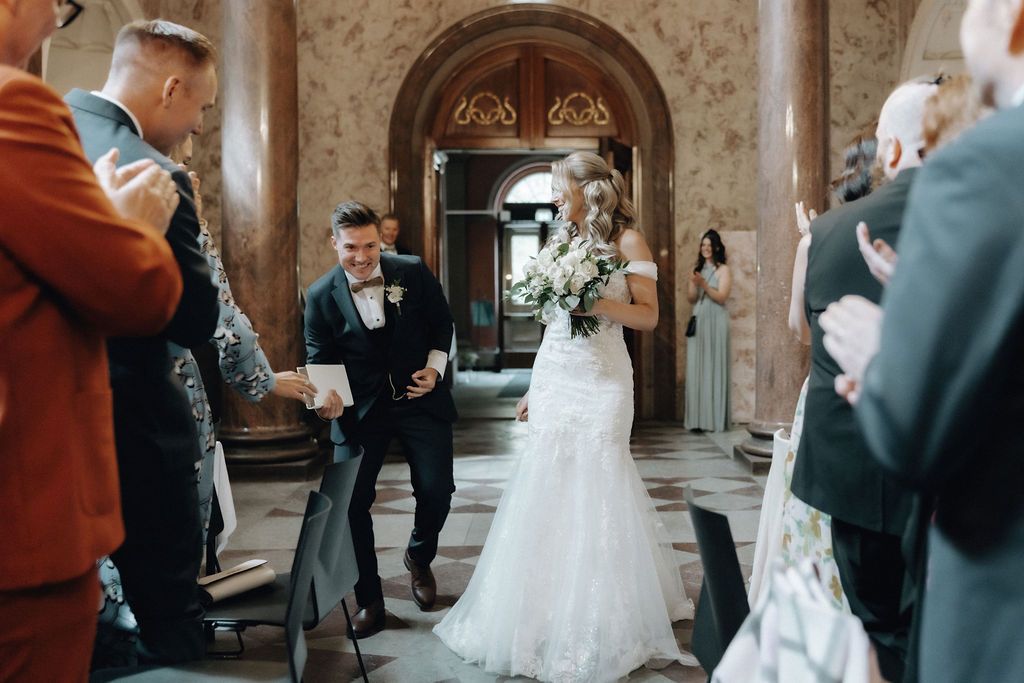 Smiling bride and groom walk down aisle surrounded by applauding guests in ornate venue.
