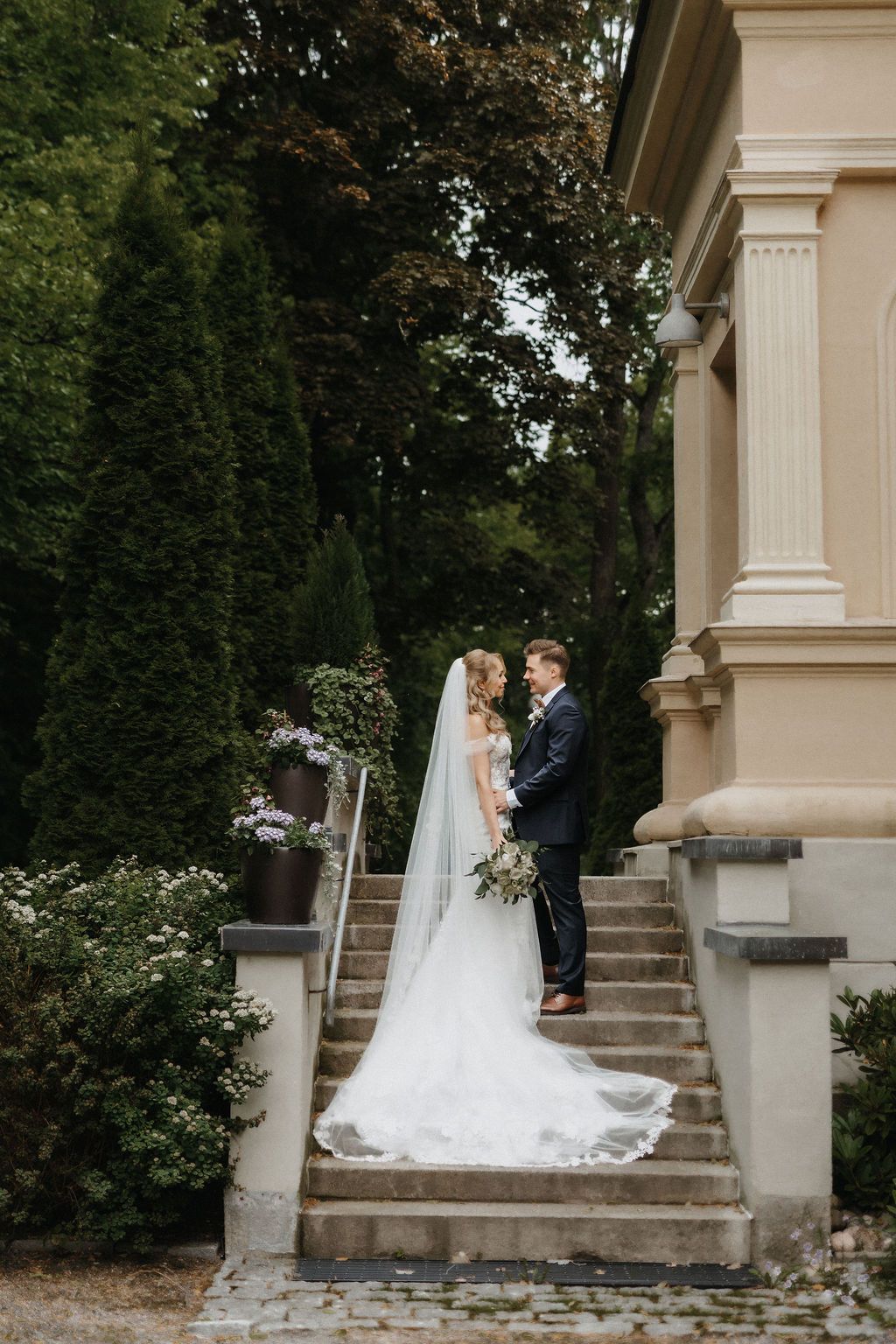 Bride and groom stand on stone steps beside elegant building surrounded by lush greenery during wedding portraits.