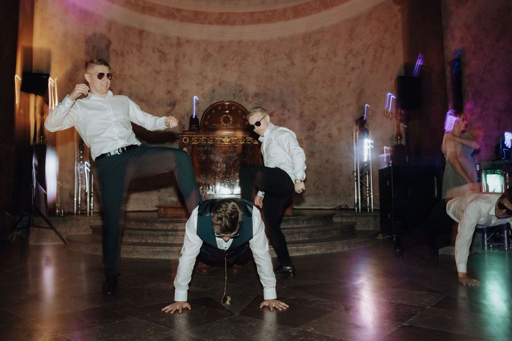 Groomsmen in sunglasses performing breakdance moves at a wedding reception in an elegant venue.