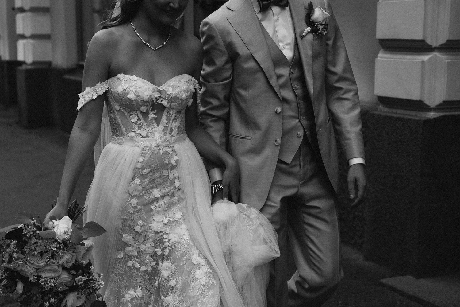 Bride and groom walk together in black and white photo, bride wearing floral off-shoulder gown.