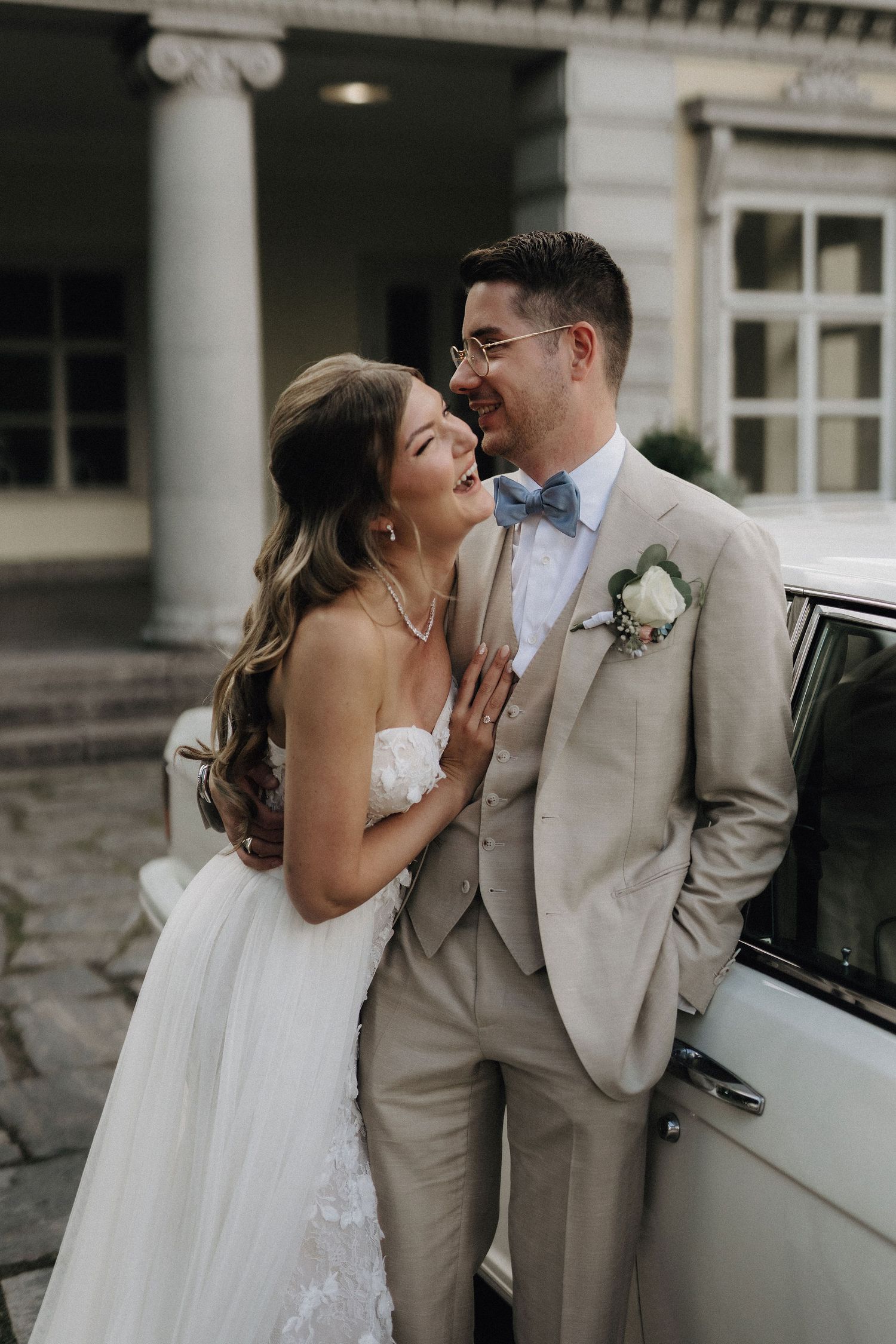Smiling bride and groom in beige suit with blue bow tie laughing together beside a vintage white car.
