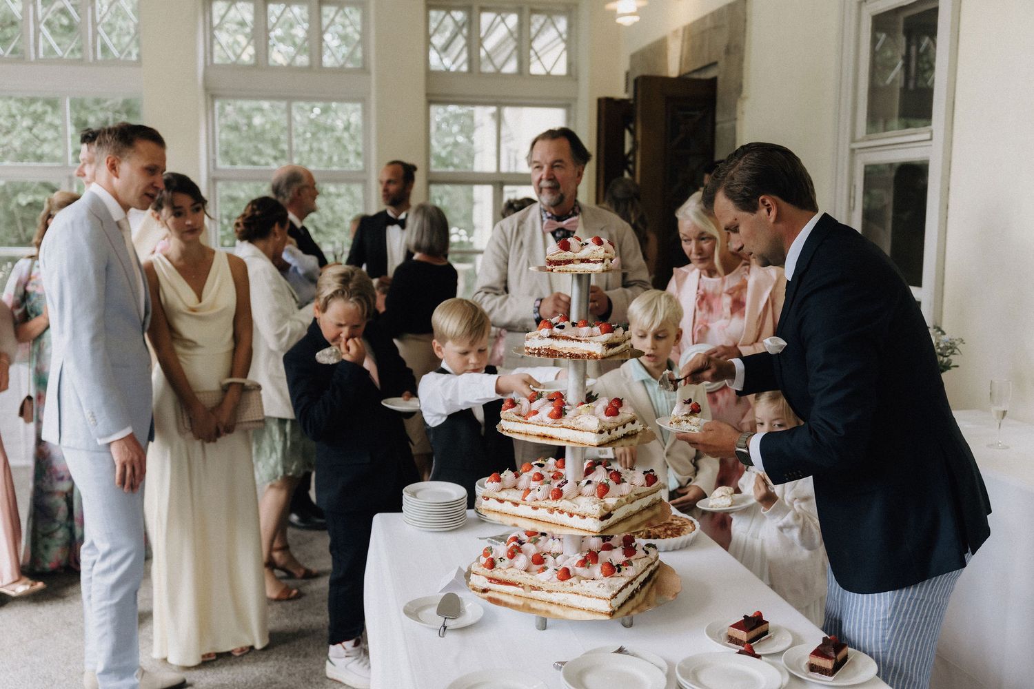 Wedding guests gathering around a tiered strawberry cake table in an elegant bright reception hall.