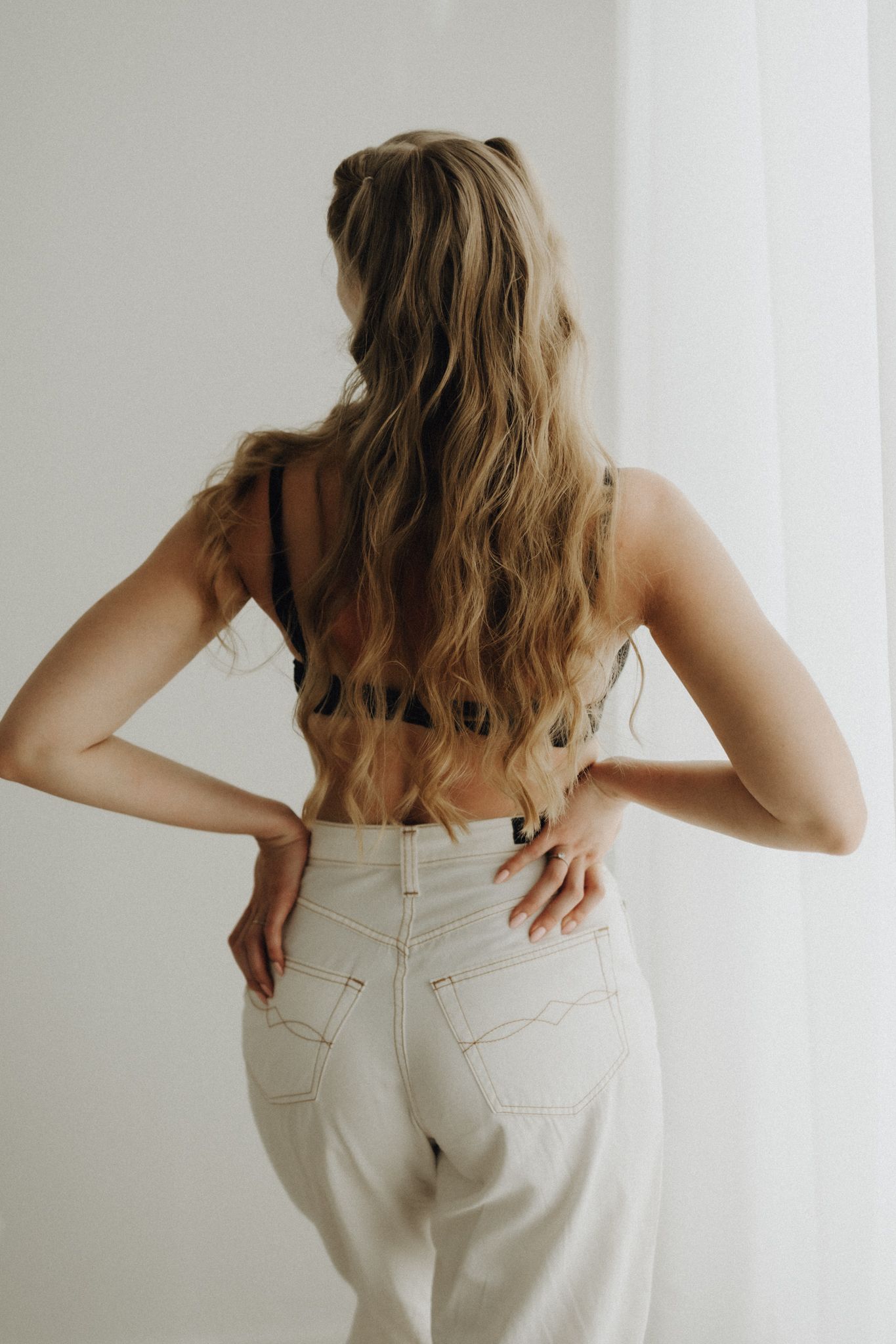 Rear view of woman with long wavy hair wearing crop top and white high-waist jeans.