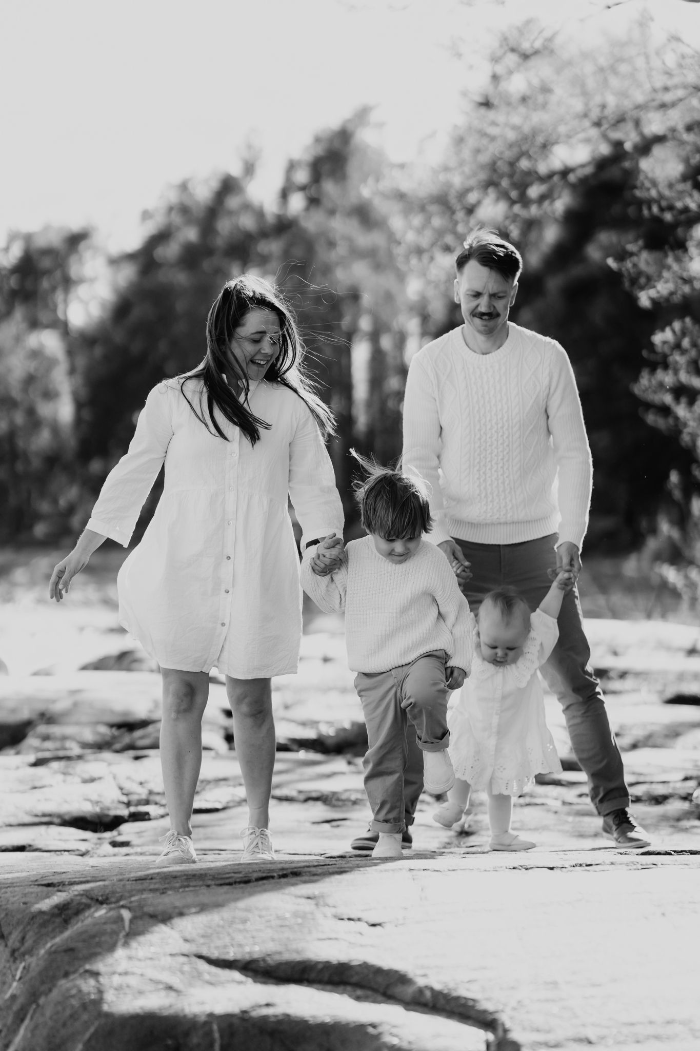 A black and white photo of a cheerful family walking hand in hand along a rocky beach with two children.