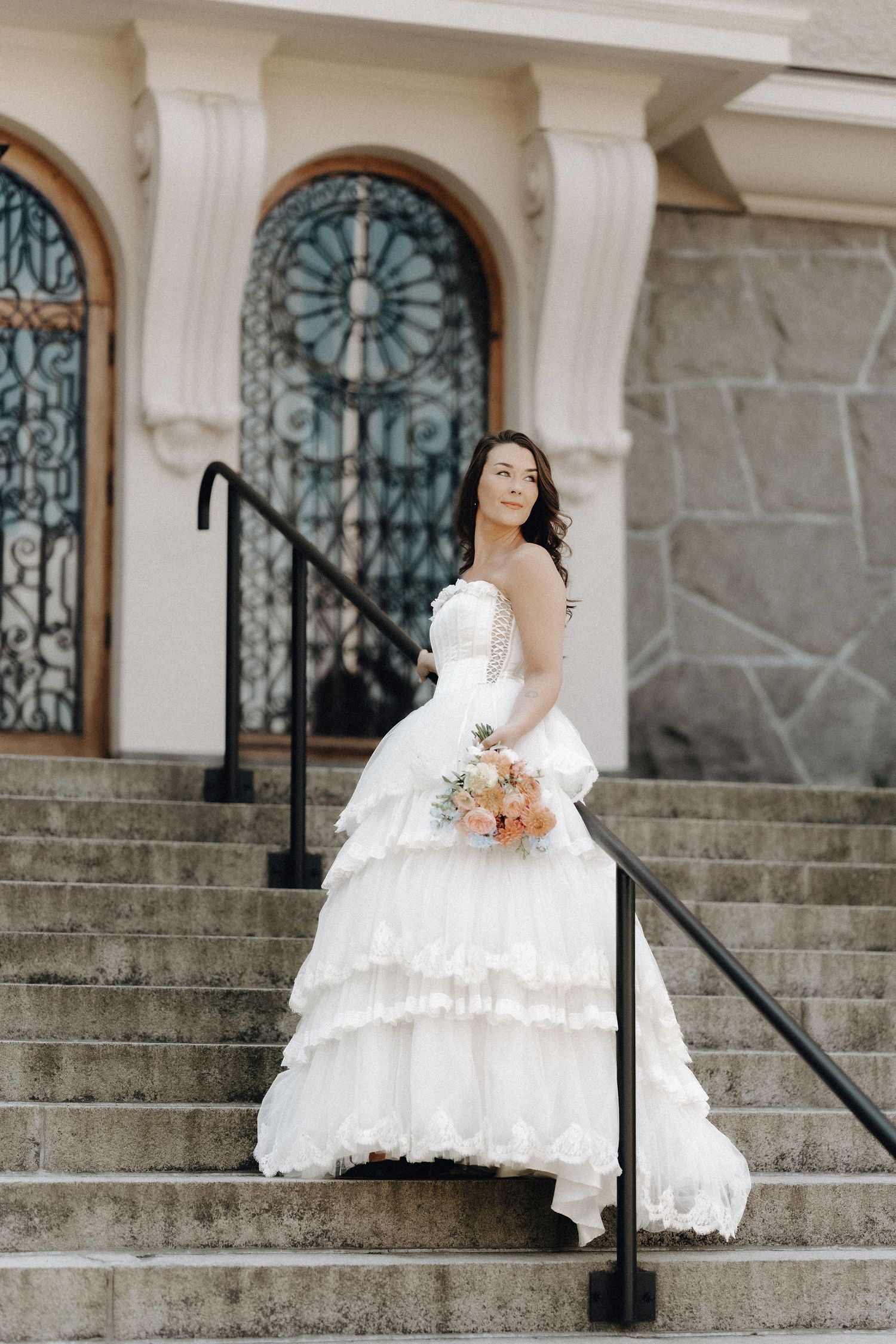 Bride in tiered white gown holding bouquet on stone steps before ornate arched iron door.