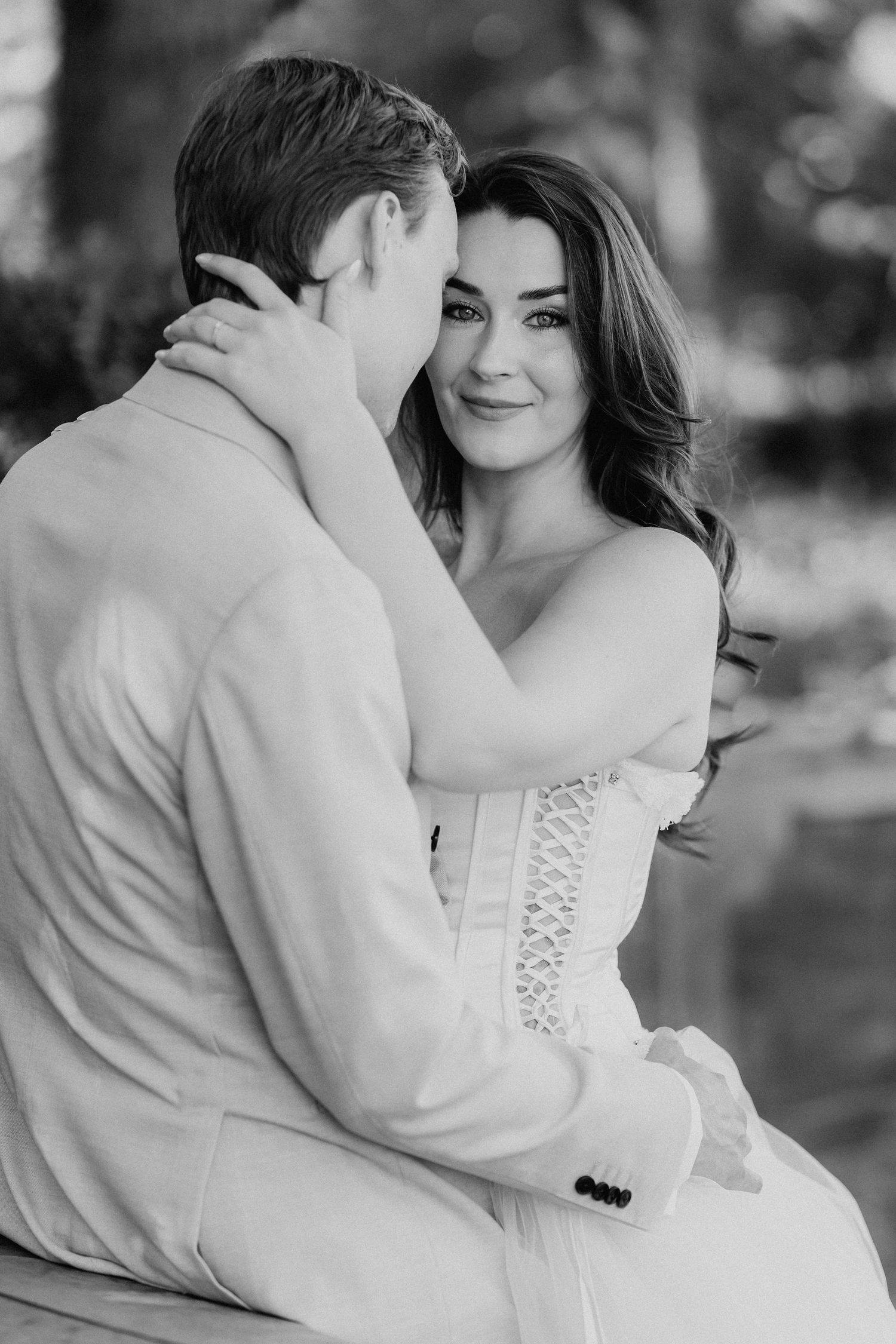 Black and white photo of bride and groom embracing tenderly outdoors, bride smiling at camera.