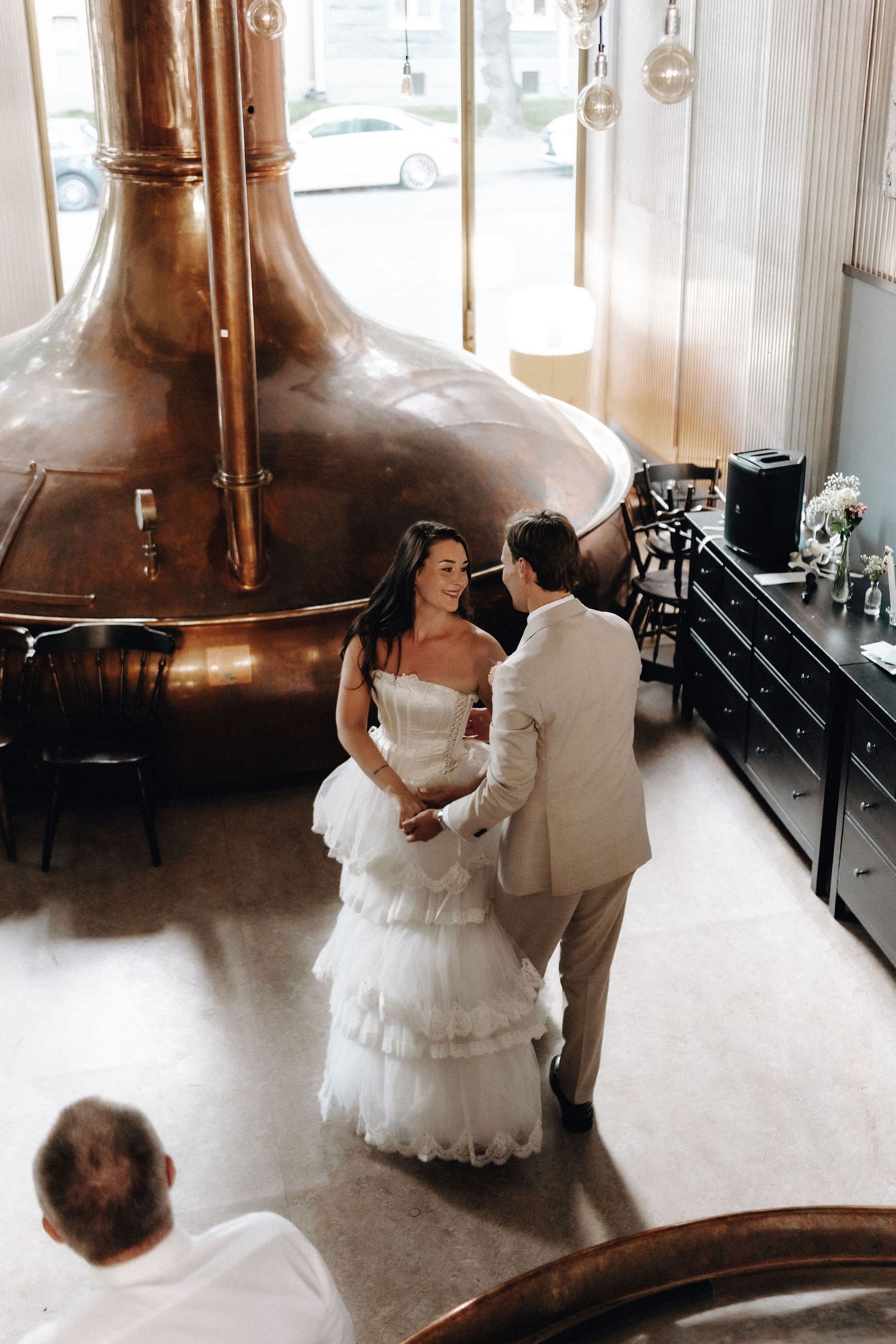 Bride and groom share an intimate moment holding hands near a large copper brewing vessel at their wedding.