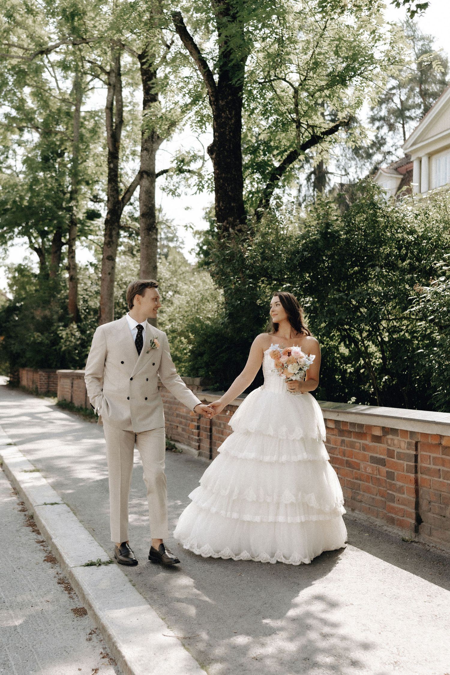 Newlyweds walk hand-in-hand on a tree-lined path, bride in tiered white gown holding a bouquet.