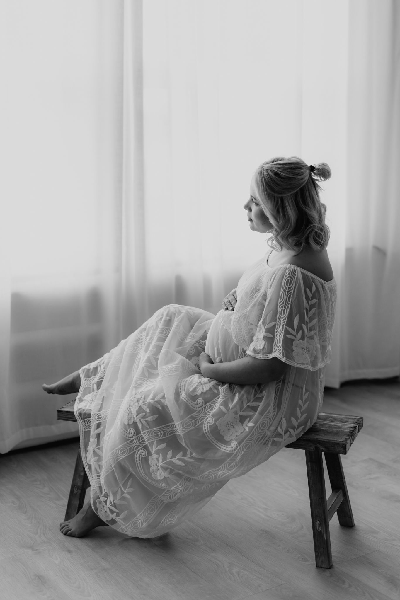 Pregnant woman in elegant white lace dress sits on wooden bench gazing toward bright window.