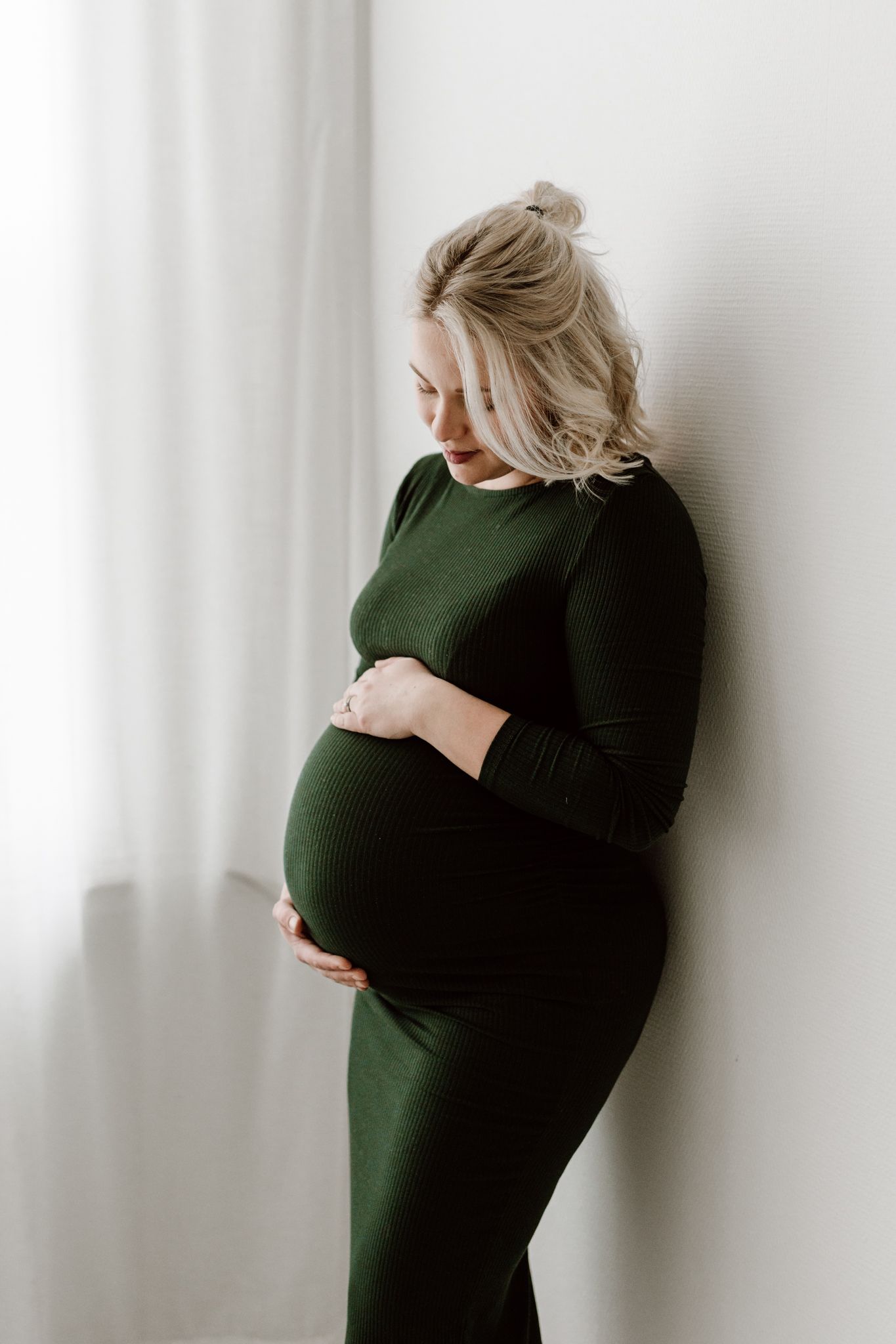 Pregnant woman in dark green long-sleeve dress gently holding baby bump near sheer curtains.