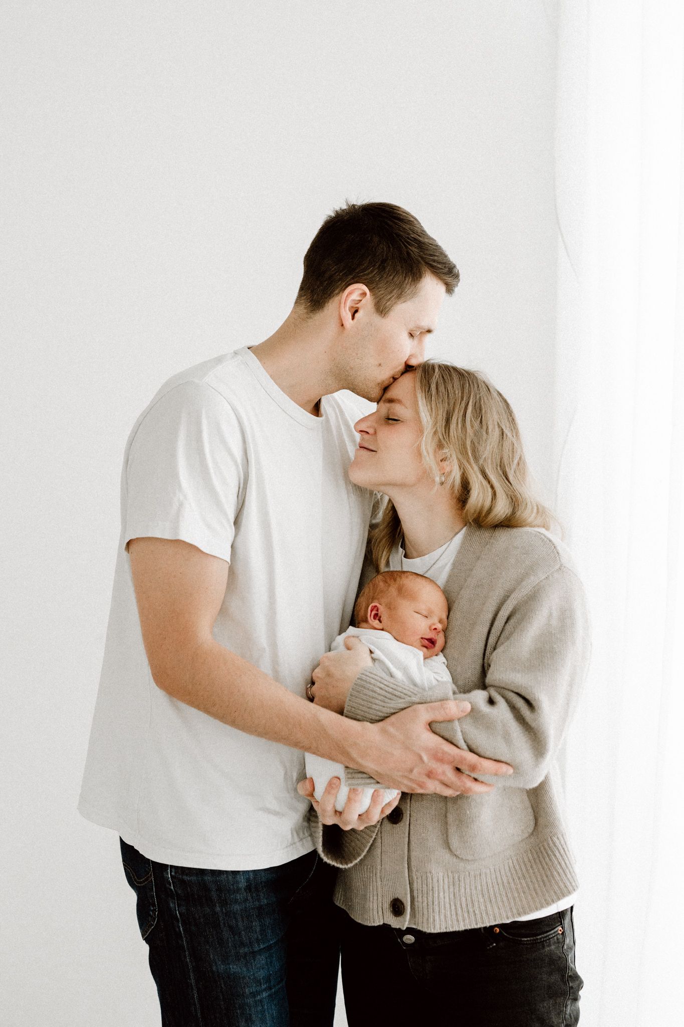 Father kissing mothers forehead as they both cradle their newborn baby against white background.