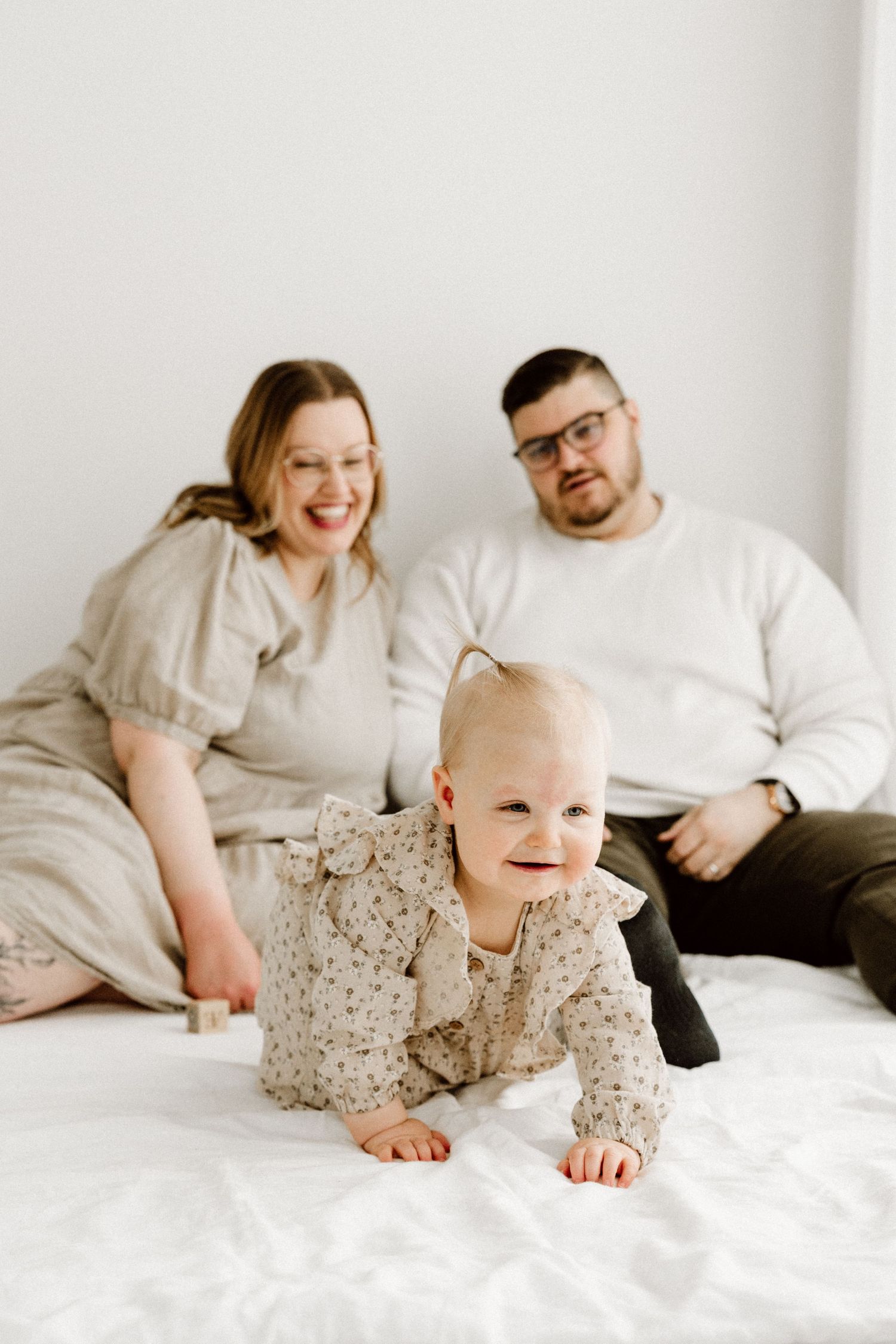 A smiling baby girl crawls on a white blanket while her parents sit and watch in a bright studio.