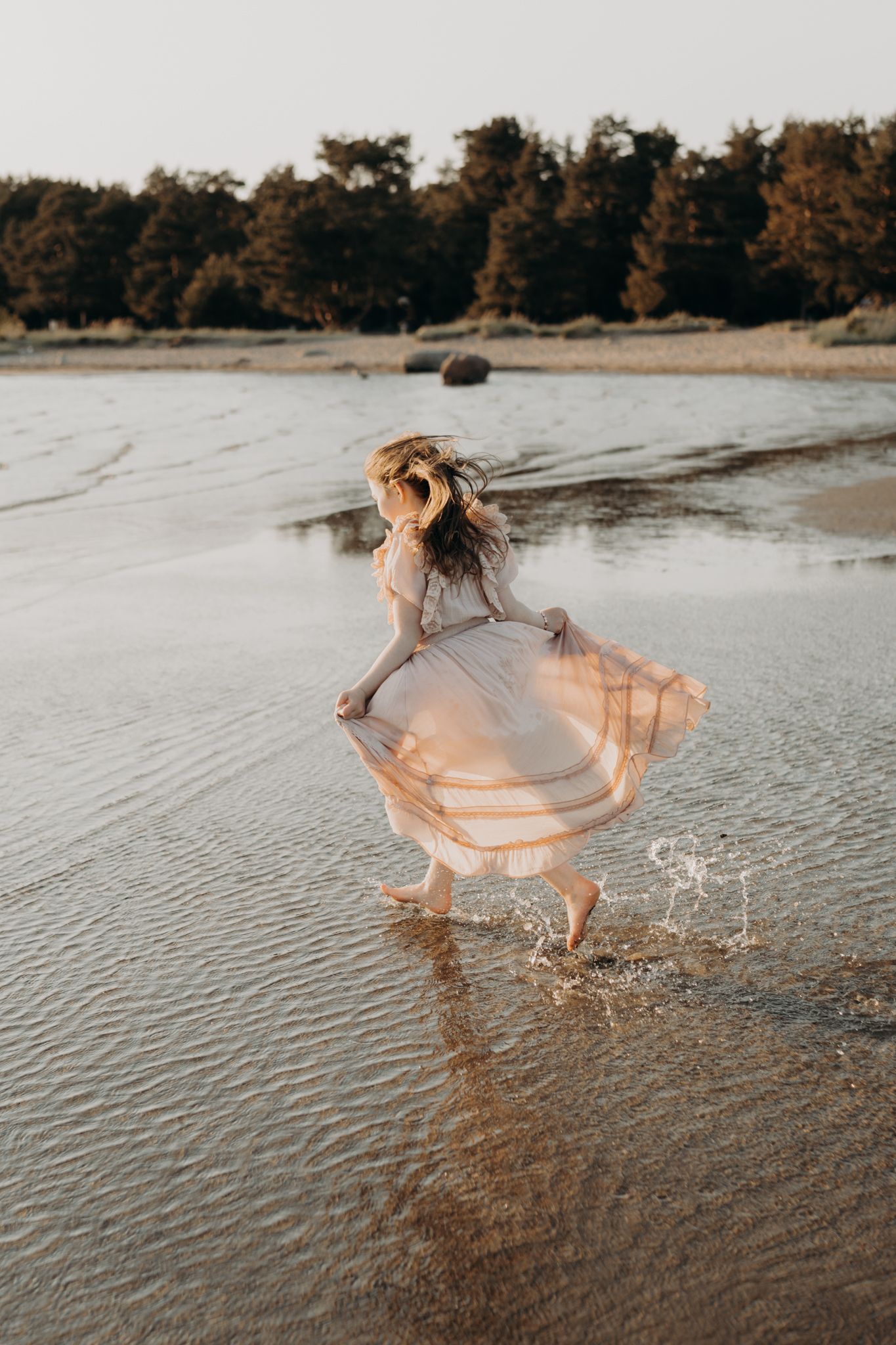 A young girl in a flowing blush dress runs barefoot through shallow water on a sunlit beach.