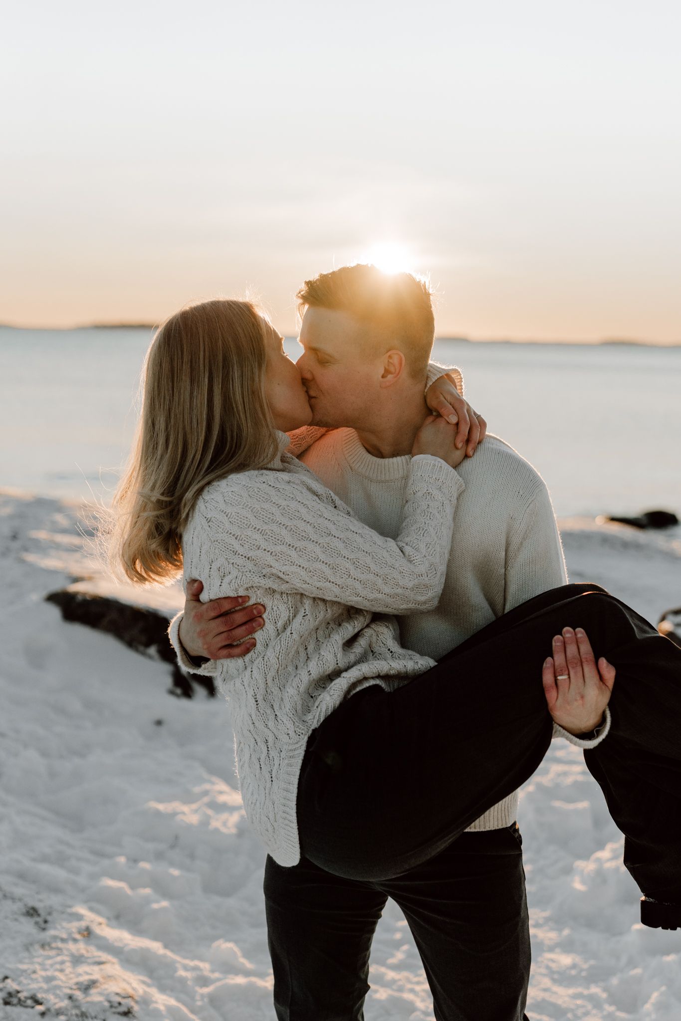 Couple shares a romantic kiss as man lifts woman on a snow-covered beach at golden sunset.