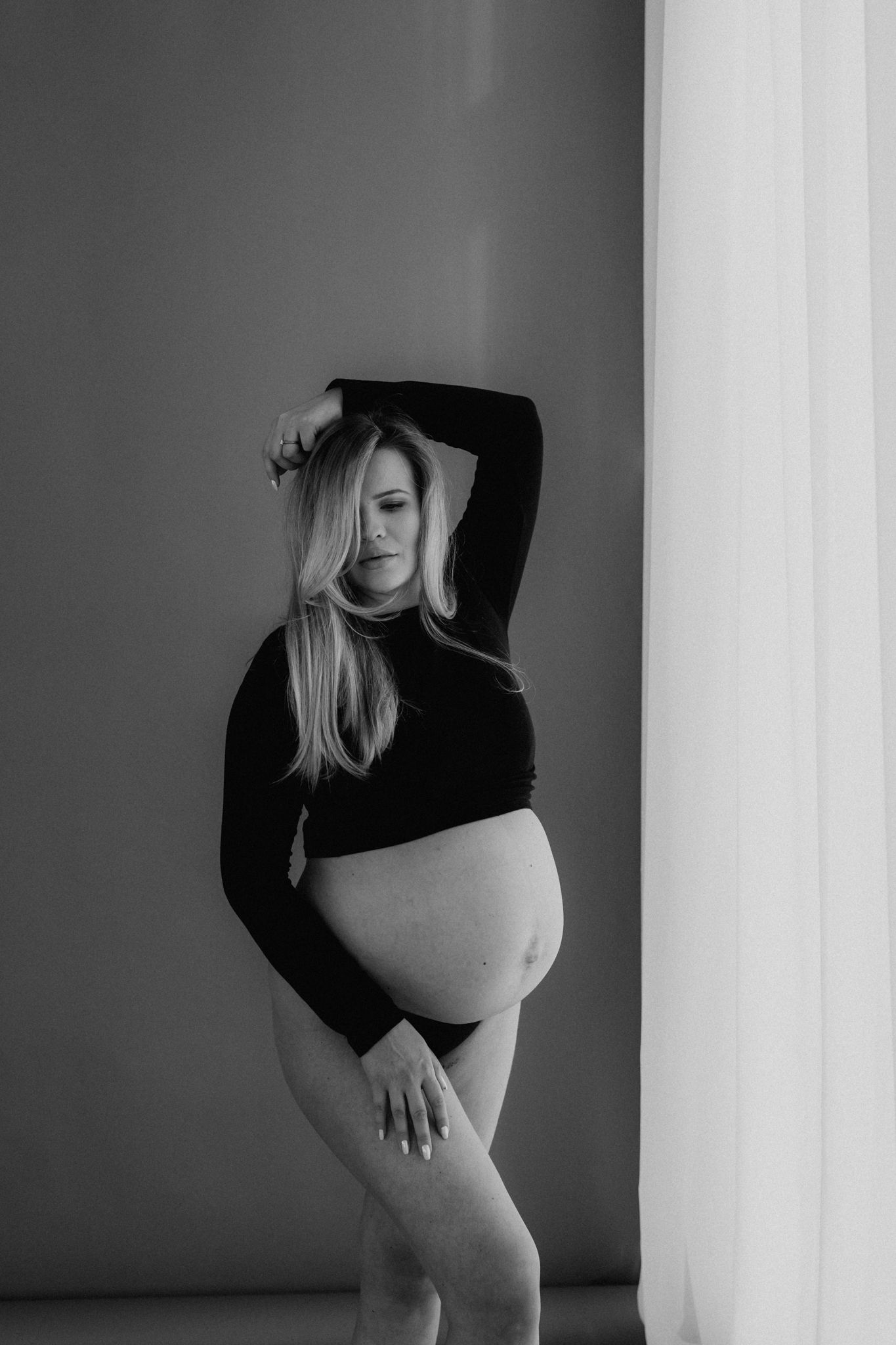 Black and white maternity portrait of pregnant woman in black outfit cradling bump near sheer curtain.